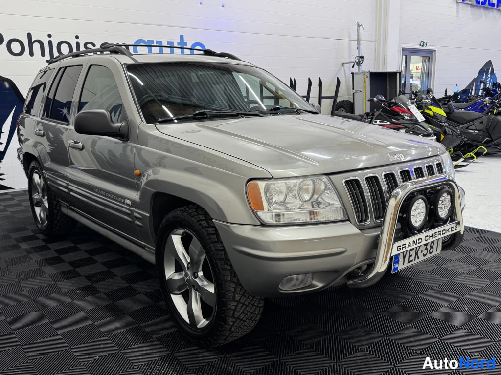 Etuviisto, nokka oikealle: Hopeanharmaa Jeep Grand Cherokee sisätilassa, etukulma oikealta; konepellillä Jeep-merkki, keulakaarella kaksi lisävaloa ja rekisterikilpi.
