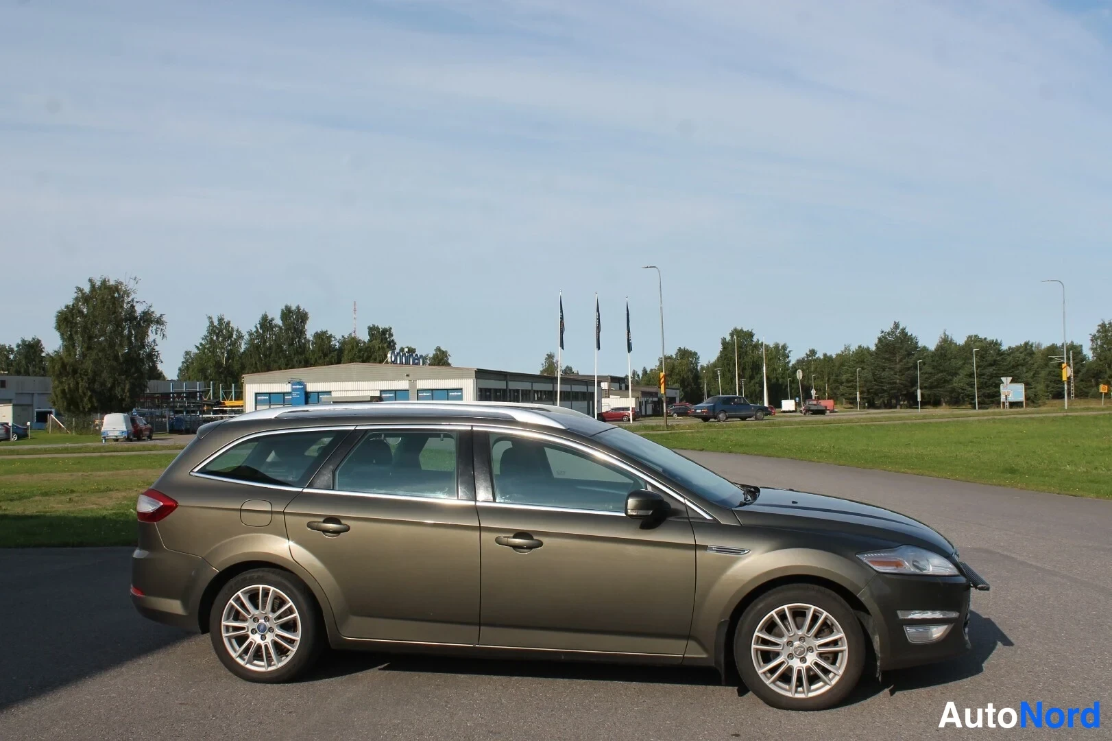Etuviisto, nokka oikealle: Farmariauto taaksepäin viistosta sivulta vasemmalle kuvattuna; tummanharmaa/oliivinsävyinen kori, kattokaiteet ja monisäteittäiset alumiinivanteet näkyvä
