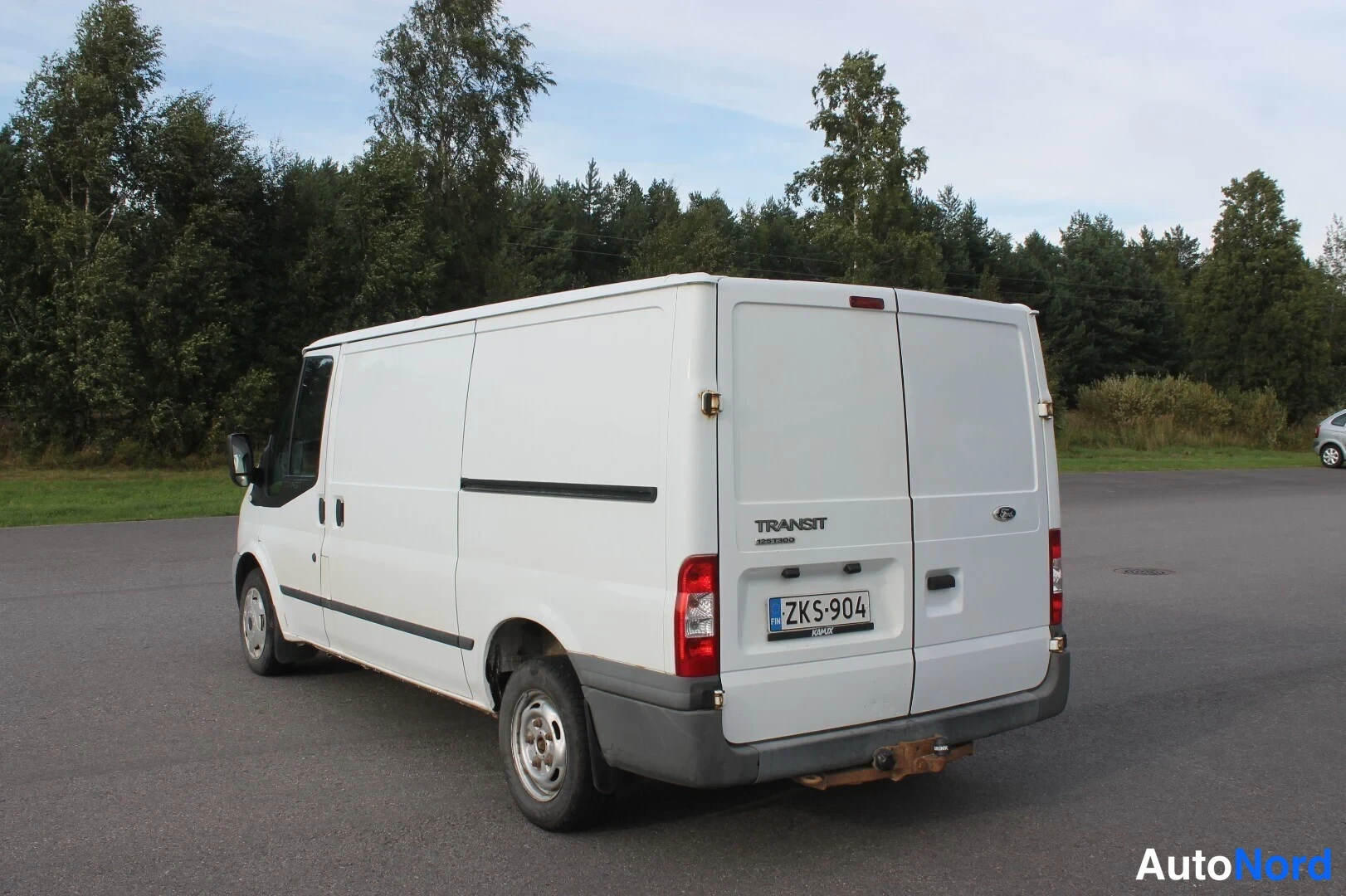Takaviisto: Valkoinen Ford Transit -pakettiauto takaa ja vasemmalta kyljestä kuvattuna parkkipaikalla metsämaiseman edessä. Takapenkin rekisteritunnus ja Transit-merkintä n