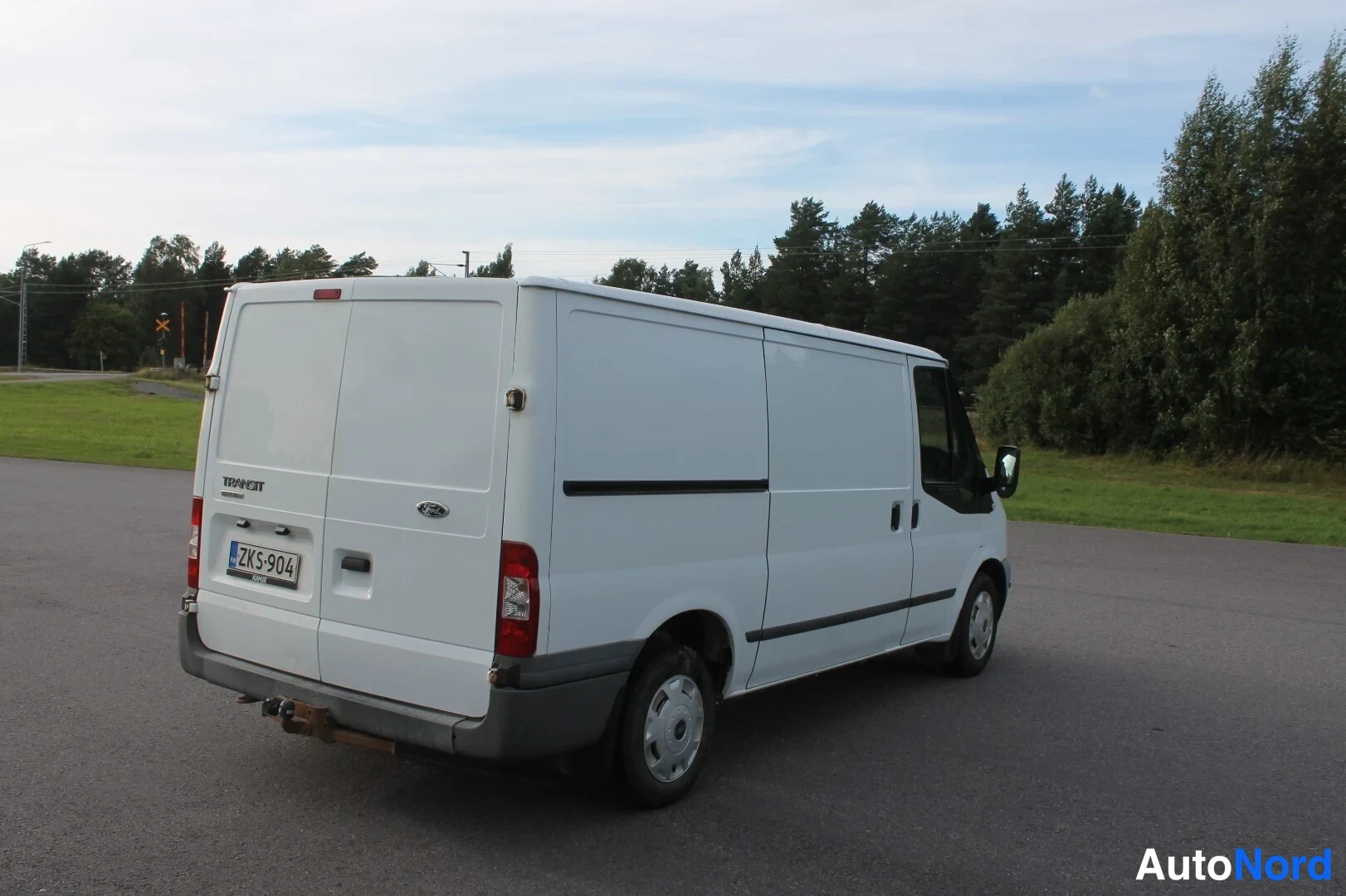 Takaviisto: Ford Transit -pakettiauton takakulman kuva: valkoinen koriovi, takavalot ja rekisterikilpi näkyvät.