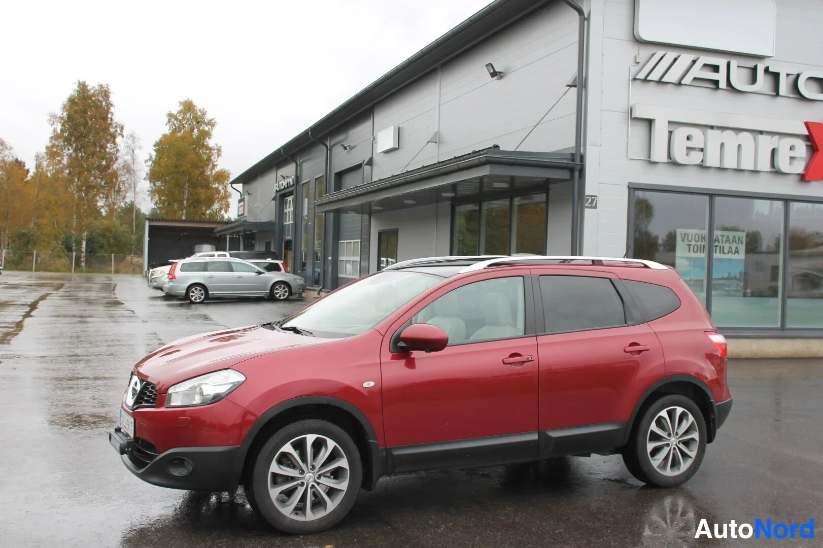 Etuviisto, nokka vasemmalle: Punainen Nissan Qashqai+2 etusivulta vasemmalta hallin edustalla, kattokaiteet ja takavalo näkyvissä.