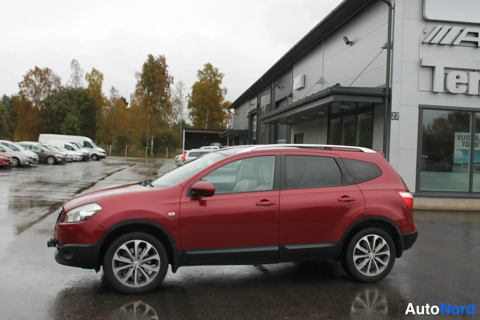 Etuviisto, nokka vasemmalle: Punainen Nissan Qashqai+2 etusivulta vasemmalta parkkipaikalla, kylki ja etuosa näkyvissä.