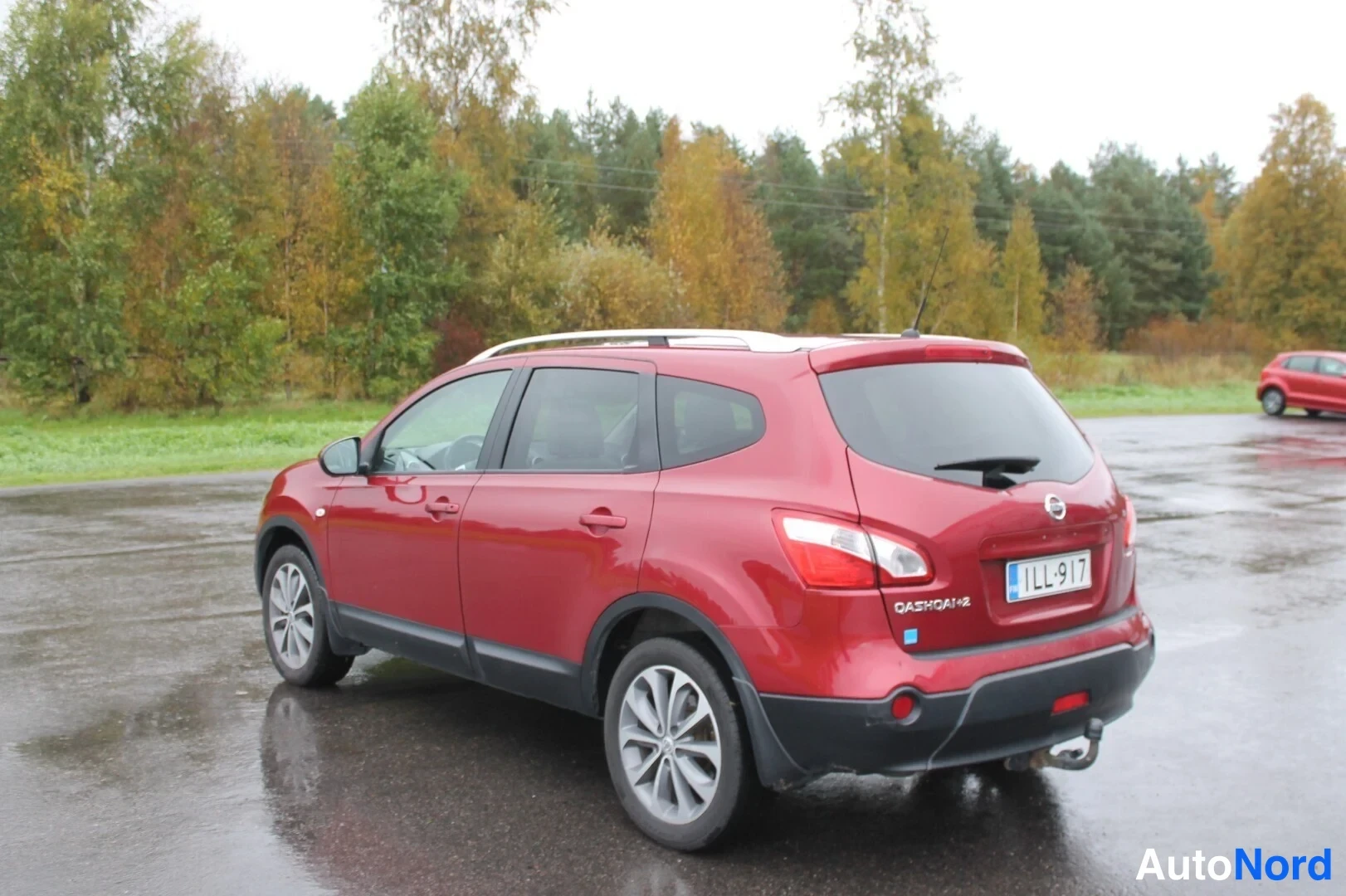Takaviisto: Punainen Nissan Qashqai+2 -maasturin takakulma märällä parkkipaikalla, rekisterikilpi, kattokaiteet ja rekaveto näkyvät.