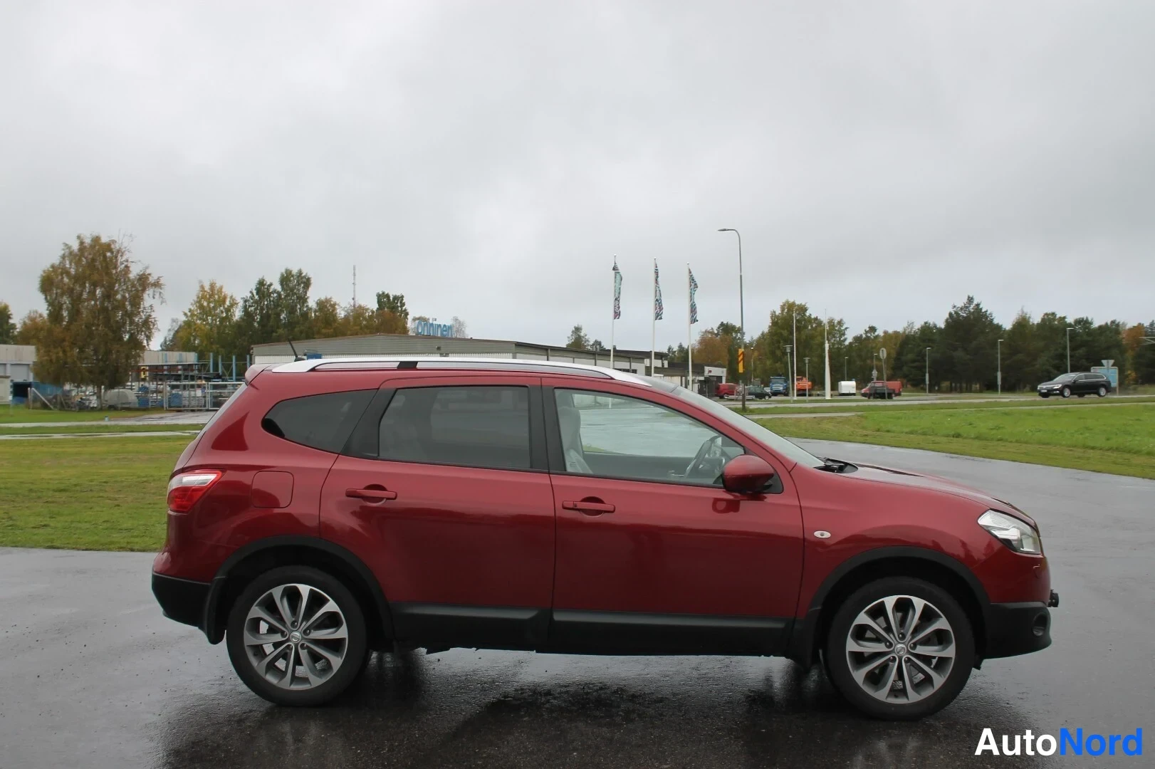 Sivulta: Punaisen Nissan Qashqai+2:n sivuprofiili parkkipaikalla, kattokaiteet näkyvät.