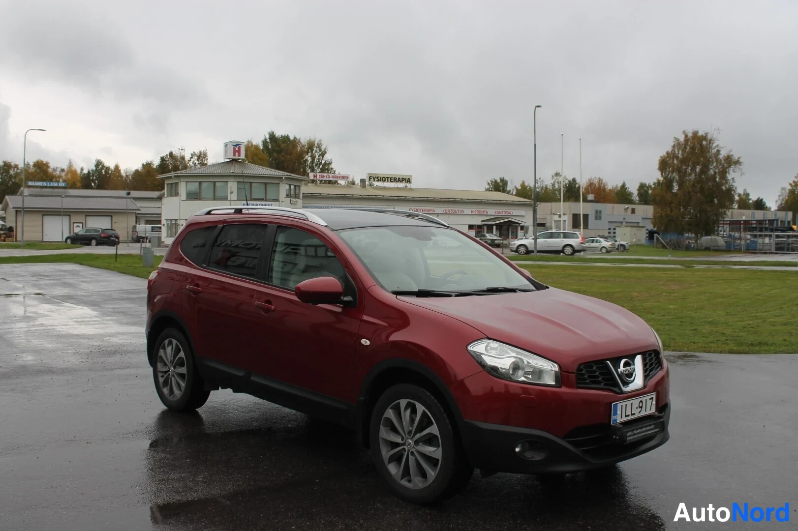 Etuviisto, nokka oikealle: Punainen Nissan Qashqai kulmassa edestä oikealta sateisella parkkipaikalla.