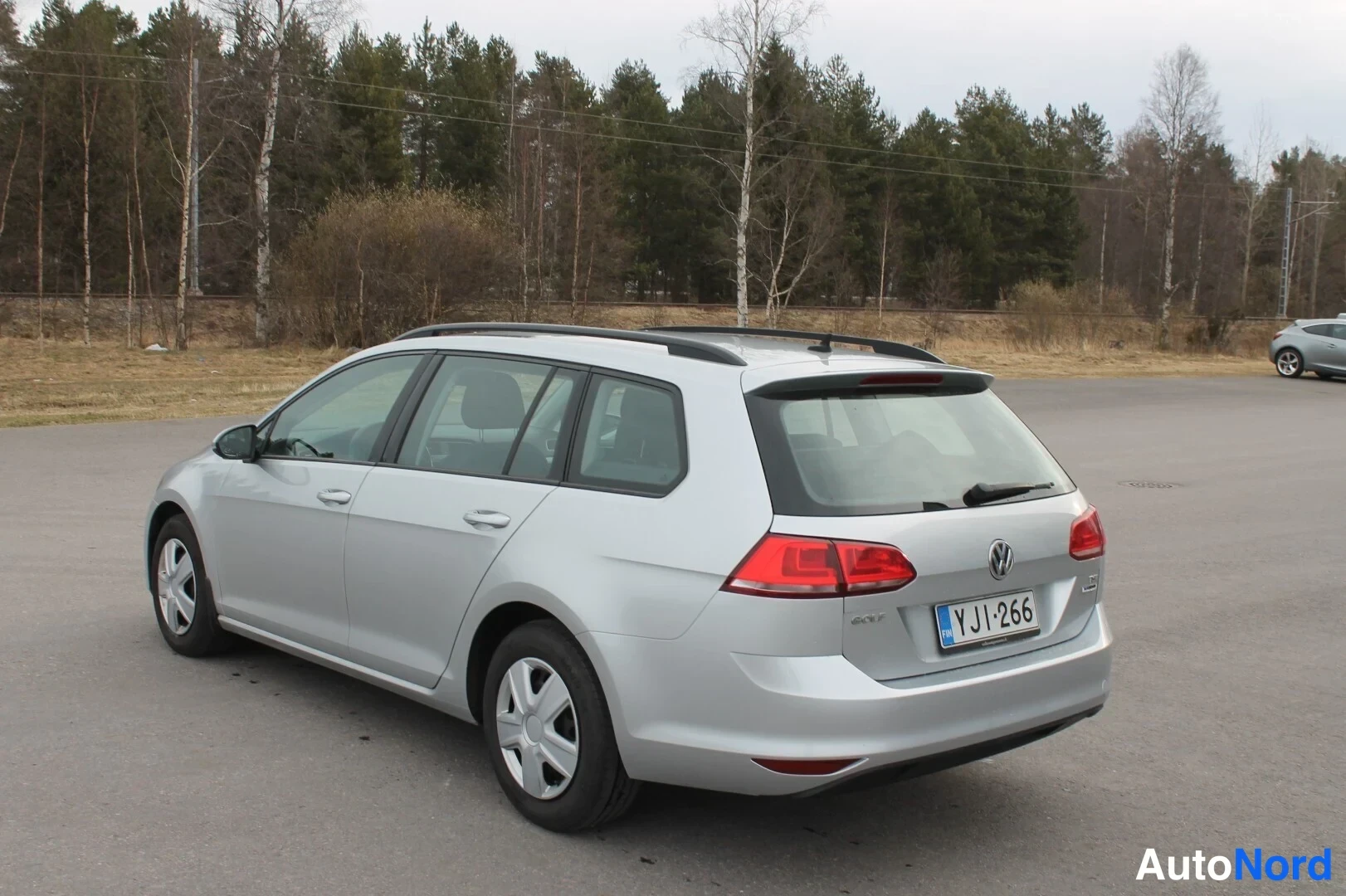 Takaviisto: Hopeinen farmariauto takaa vasemmalta kolmesta neljäsosasta, rekisterikilpi, taustalla metsää ja parkkipaikka.