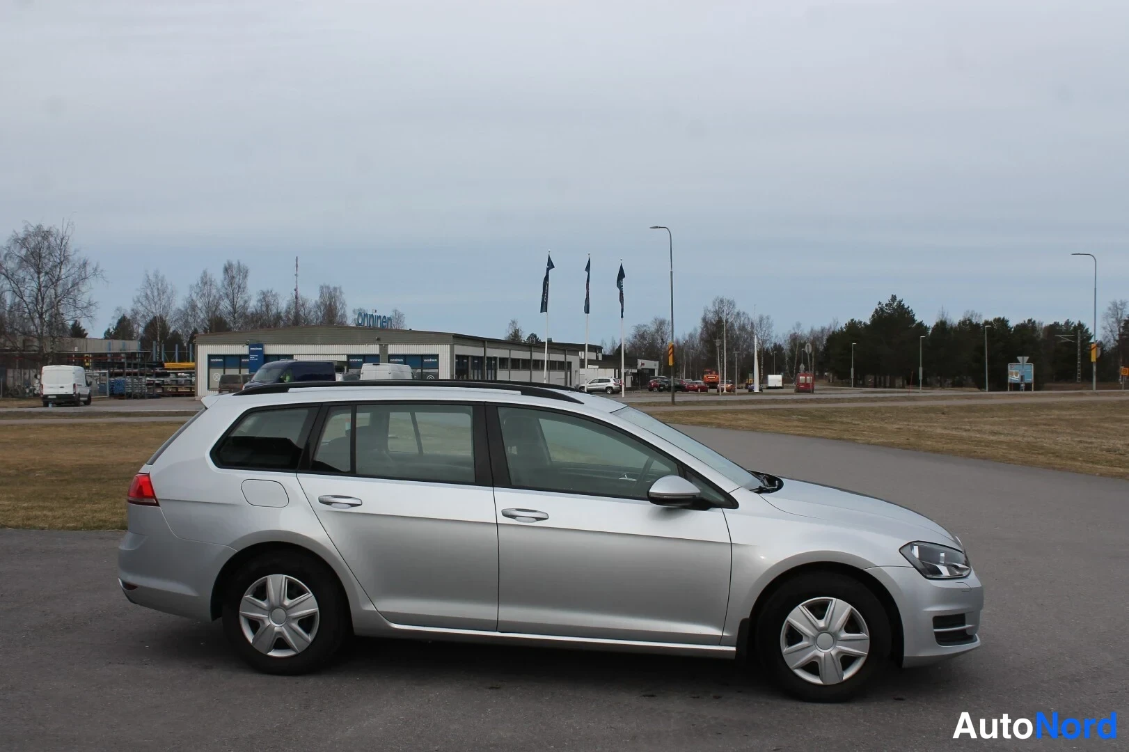 Etuviisto, nokka oikealle: Sivulta hieman viistosti kuvattu hopeinen farmariauto autopaikalla, rekisterikilpi ja etuvalo näkyvät oikealla reunalla.