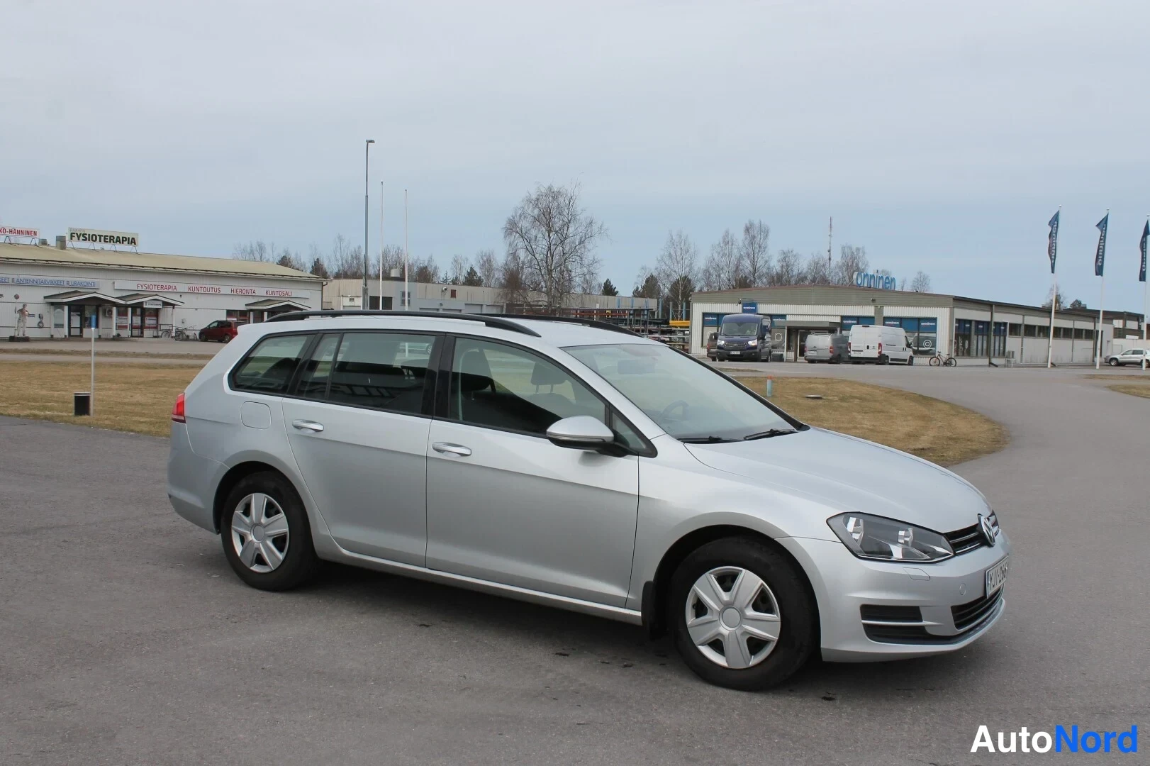 Etuviisto, nokka oikealle: Hopeinen farmariauto kuvattu etuviistosta oikealta; kylki ja keulan etuvalot näkyvät, taustalla rakennuksia ja lippuja.