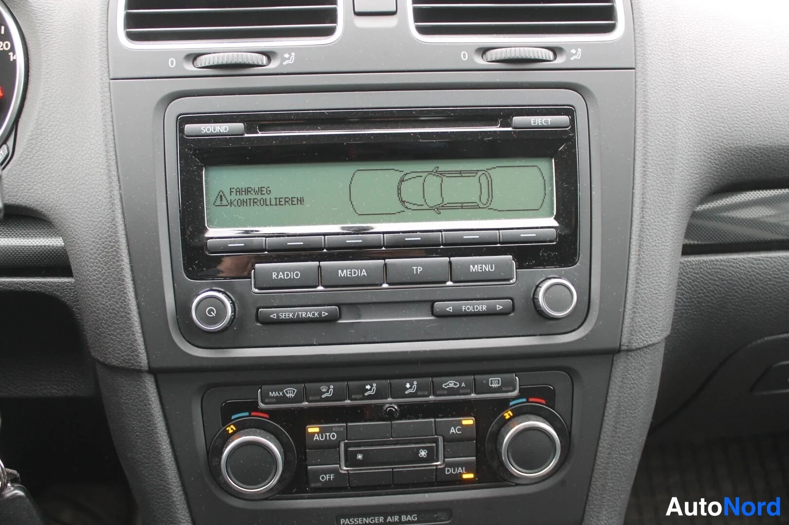 Muu: Keskikonsolin lähikuva: RCD-radio-näyttö tekstillä 'FAHRWEG KONTROLLIEREN!', näppäimet RADIO, MEDIA, TP, MENU sekä ilmastointipaneeli AUTO-, AC- ja DUAL-merkinn