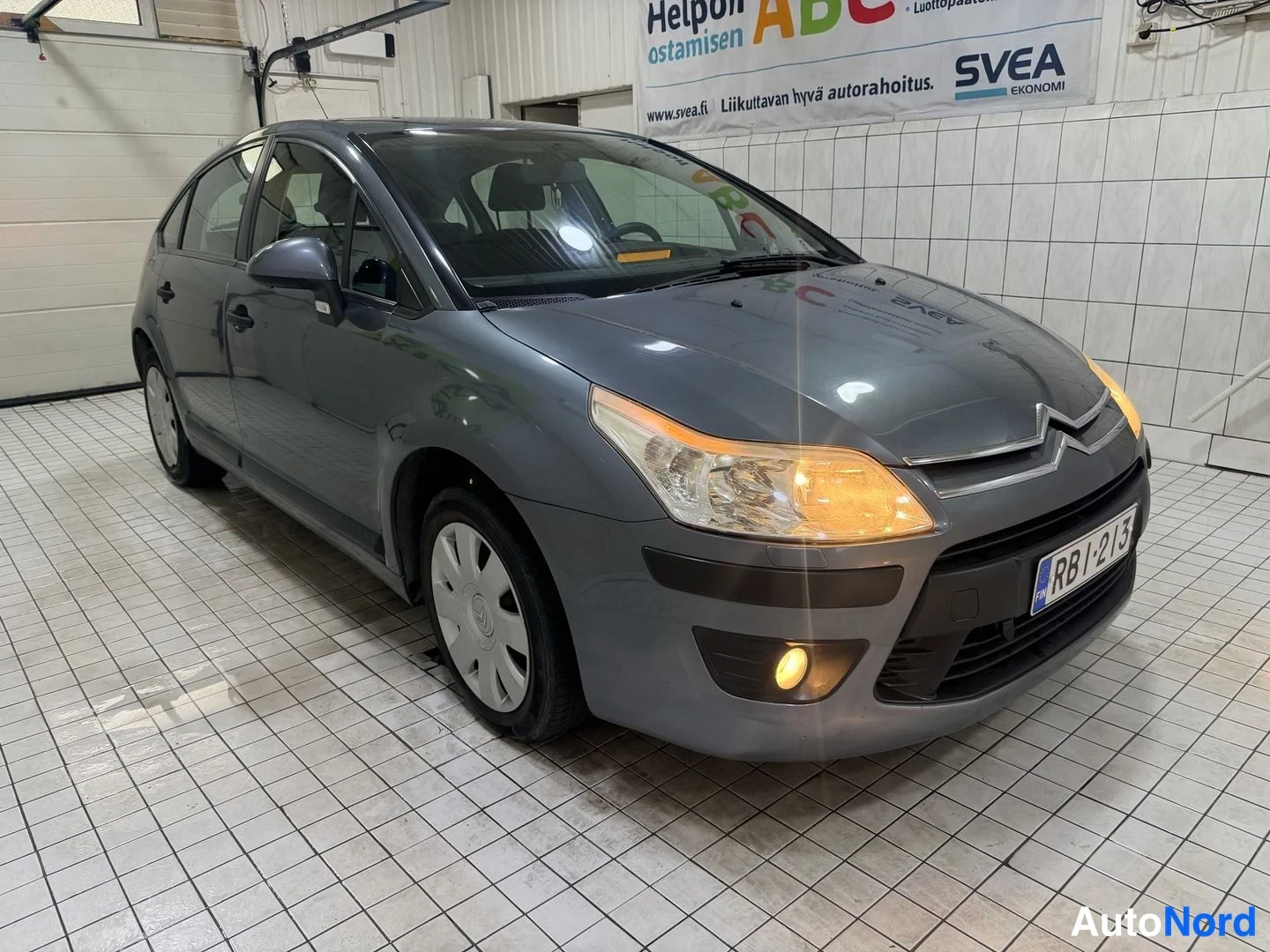 Etuviisto, nokka oikealle: Harmaa Citroën-hatchback etuviistosta; rekisterikilpi RB1-213, auto sisätilassa valkoisella laatoitetulla lattialla