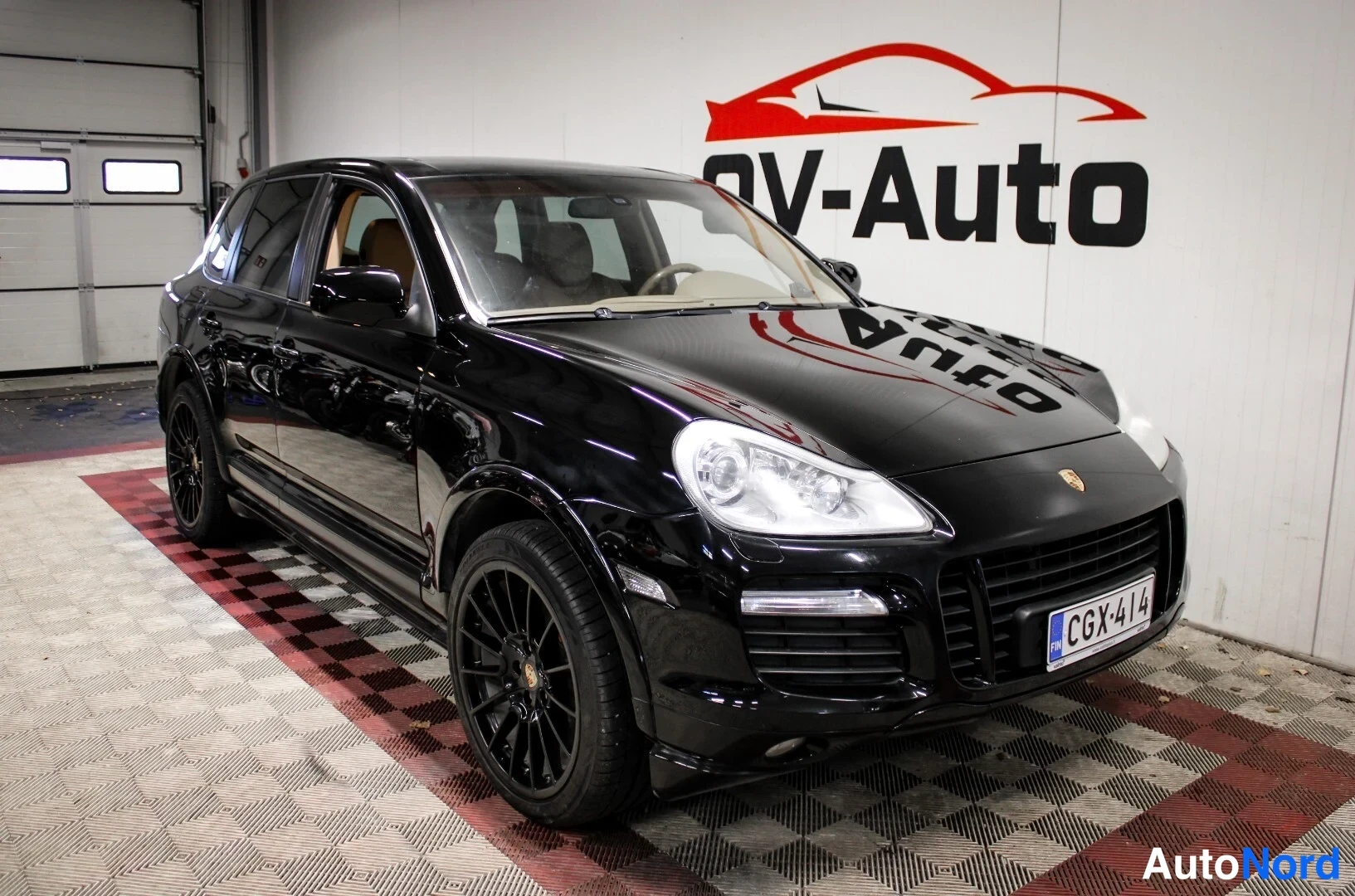 Etuviisto, nokka oikealle: Mustan Porsche Cayennen etukulmanäkymä, tummat kevytmetallivanteet ja rekisteritunnus, OV-Auton tilassa.