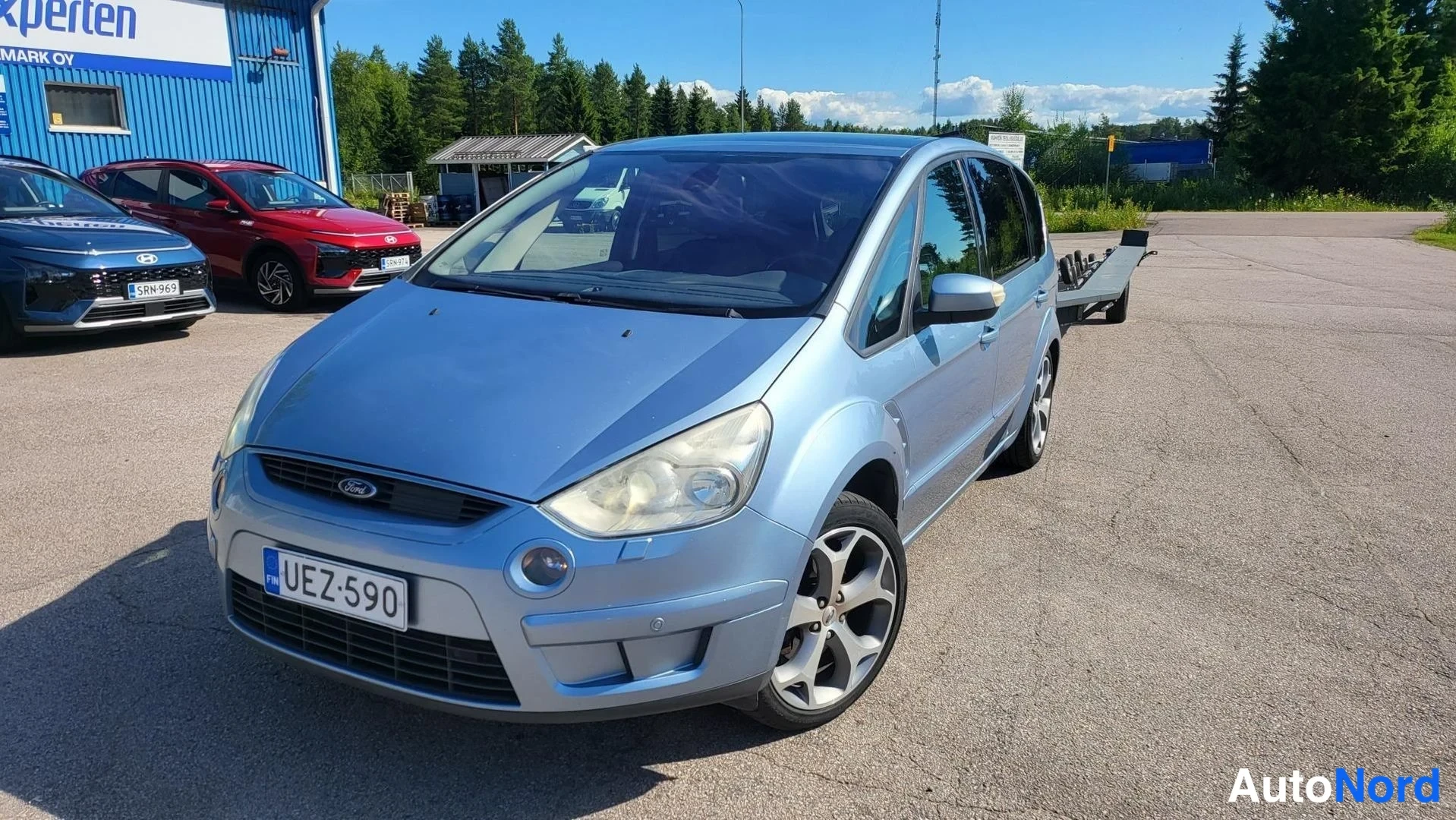 Etuviisto, nokka vasemmalle: Sininen Ford S-MAX kolmivartin etunäkymässä oikeasta sivusta, rekisteritunnus, autokuljetusperävaunu taustalla.
