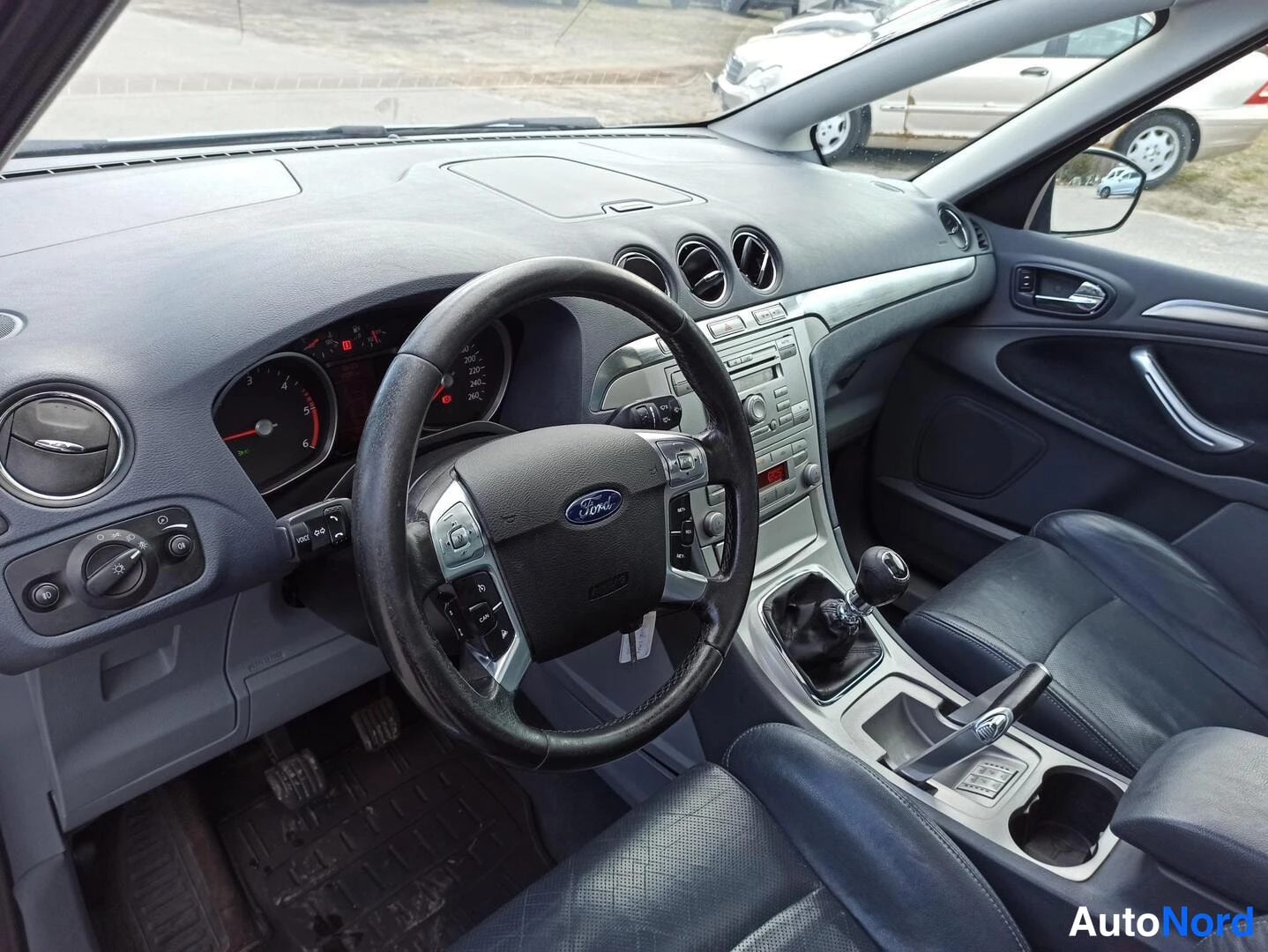 Muu: Ford S-MAXin ohjaamo: tumma kangaspenkki, Ford-merkkinen ratto, manuaalivaihteisto ja keskikonsoli näkyvät.