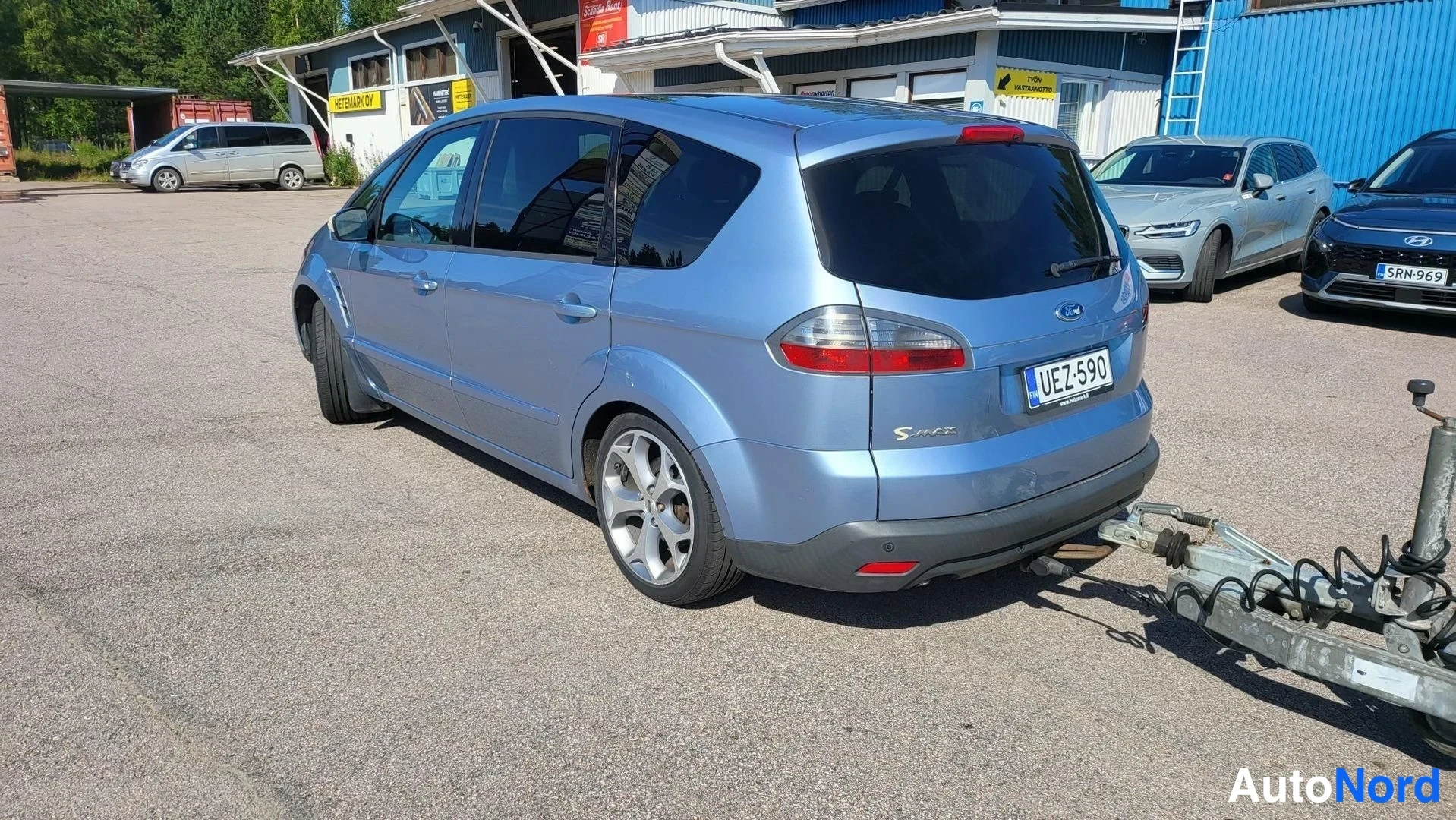 Takaviisto: Vaaleansininen tila-auto takaviistossa, rekisterikilpi, perä kiinnitetty runkovaunuun.