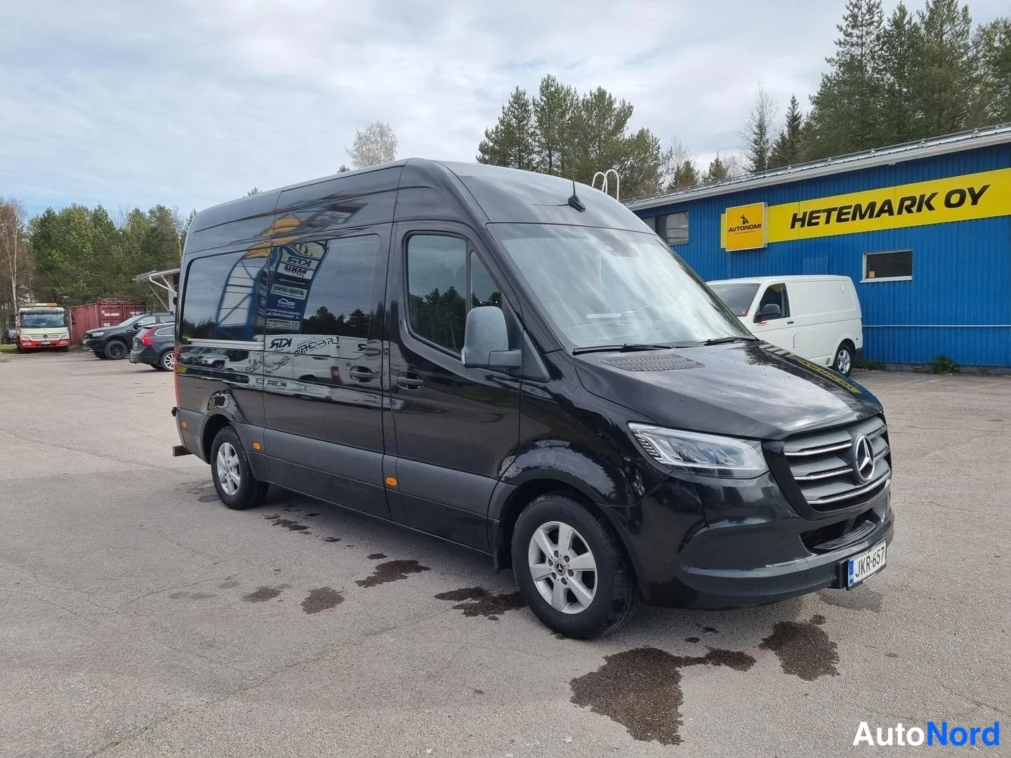 Etuviisto, nokka oikealle: Musta Mercedes-Benz Sprinter -pakettiauto kuvattu kolmivartaloasennosta edestä oikealta; konepelti, etusäleikkö ja ajovalot näkyvät.