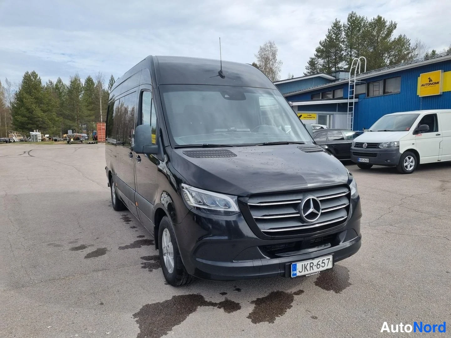 Etuviisto, nokka oikealle: Musta Mercedes-Benz Sprinter -pakettiauto kolmannelta sivulta kuvattuna, keula ja vasen kylki näkyvissä.