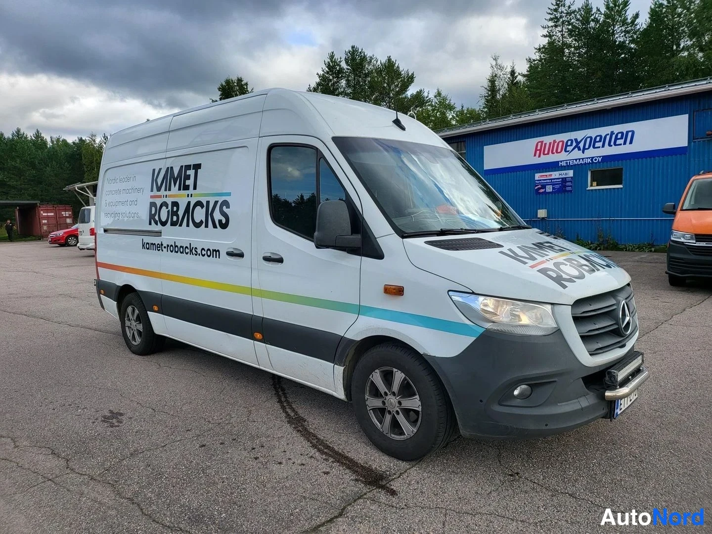 Etuviisto, nokka oikealle: Mercedes-Benz Sprinter -pakettiauto edestä vasemmalta kuvattuna, valkoinen koristekuva ja mainostekstit kyljessä.