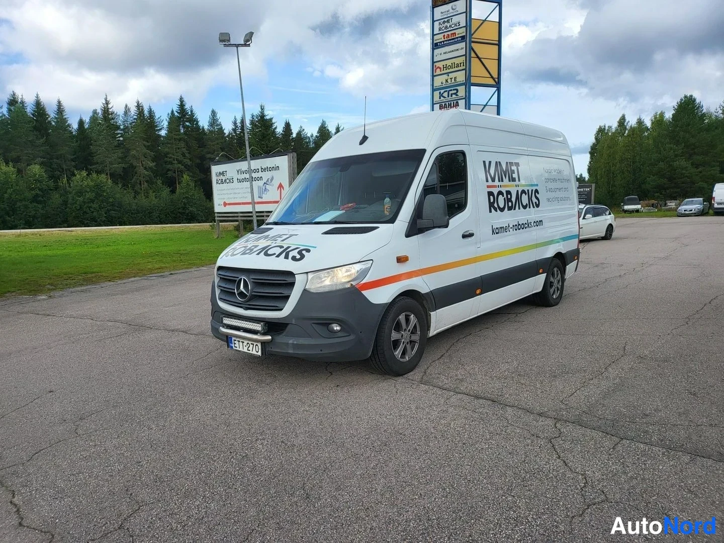 Etuviisto, nokka vasemmalle: Valkoinen Mercedes-Benz Sprinter -pakettiauto, etuosa ja vasen kylki näkyvissä, Kamet Robbacks -mainoksella.