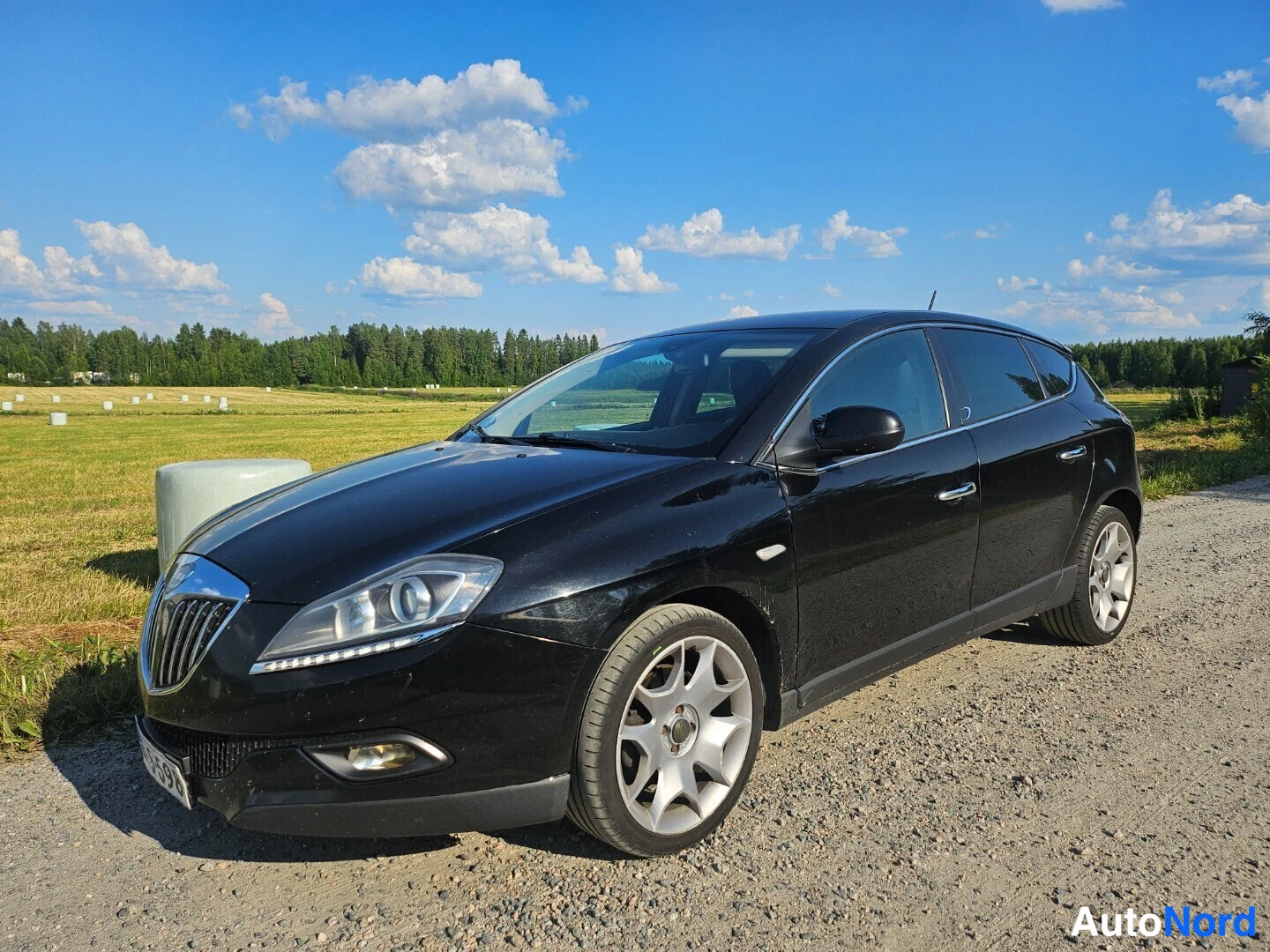 Etuviisto, nokka vasemmalle: Musta Lancia Delta kolmestinäkymä edestä vasemmalle, keula vasemmalla kuvassa soratiellä.