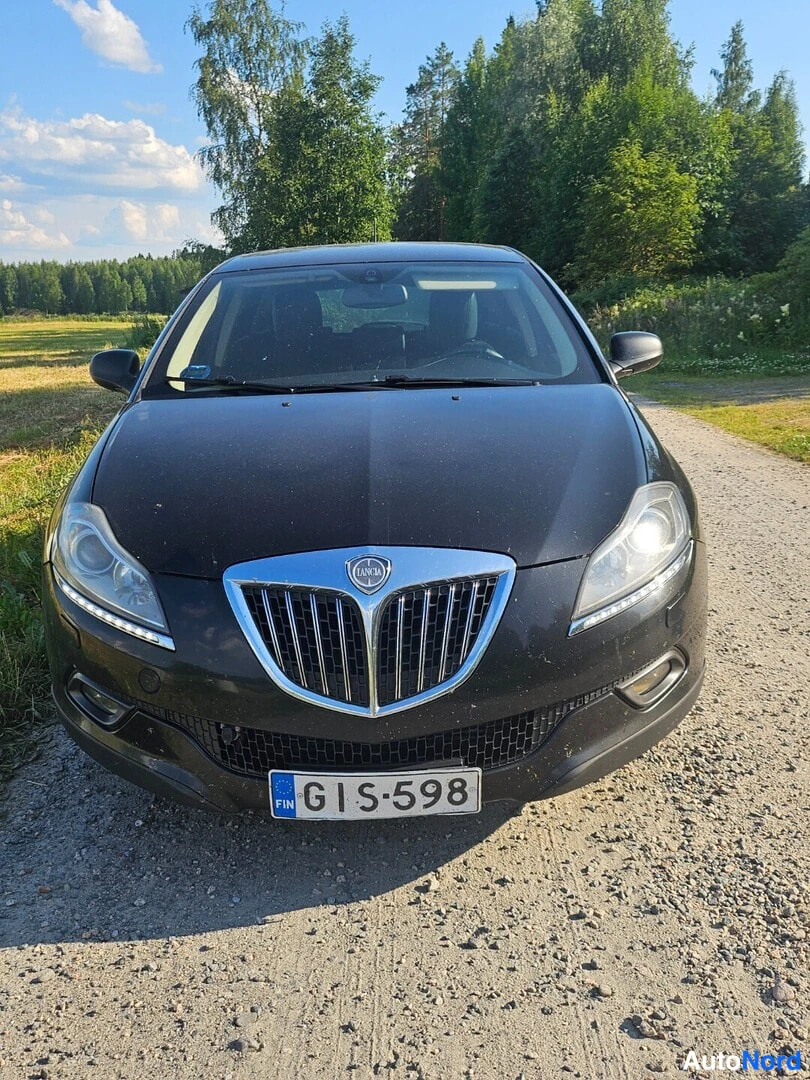 Edestä: Mustan Lancian keula suoraan edestä, rekisteritunnus, soratie ja metsä taustalla.