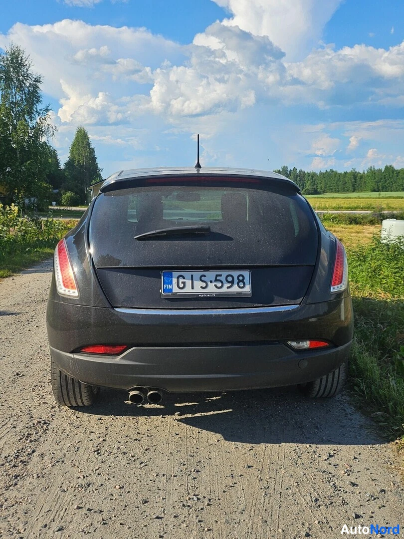Takaa: Mustan Lancian takaosa suoraan takaa, rekisteritunnus, kaksoisputkisto ja soratie.