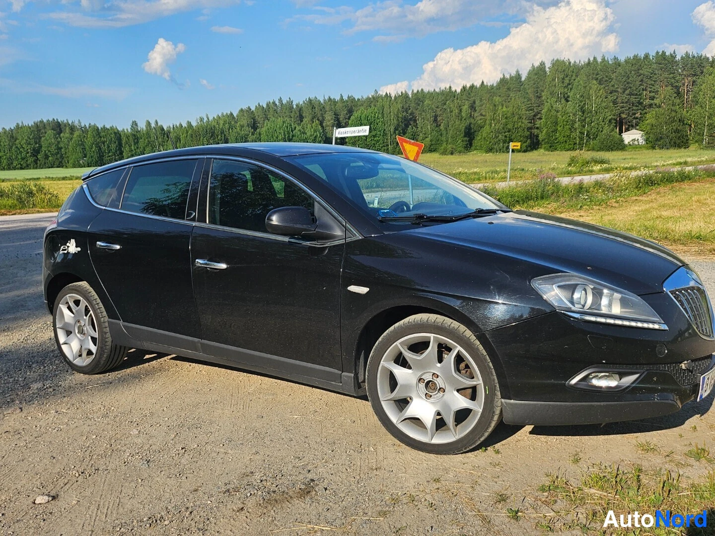 Etuviisto, nokka oikealle: Musta henkilöauto hiekkaisella pihalla, keula osoittaa oikealle; etusäleikkö ja.sumuvalot näkyvät, takana puisto ja moreeniaukko.