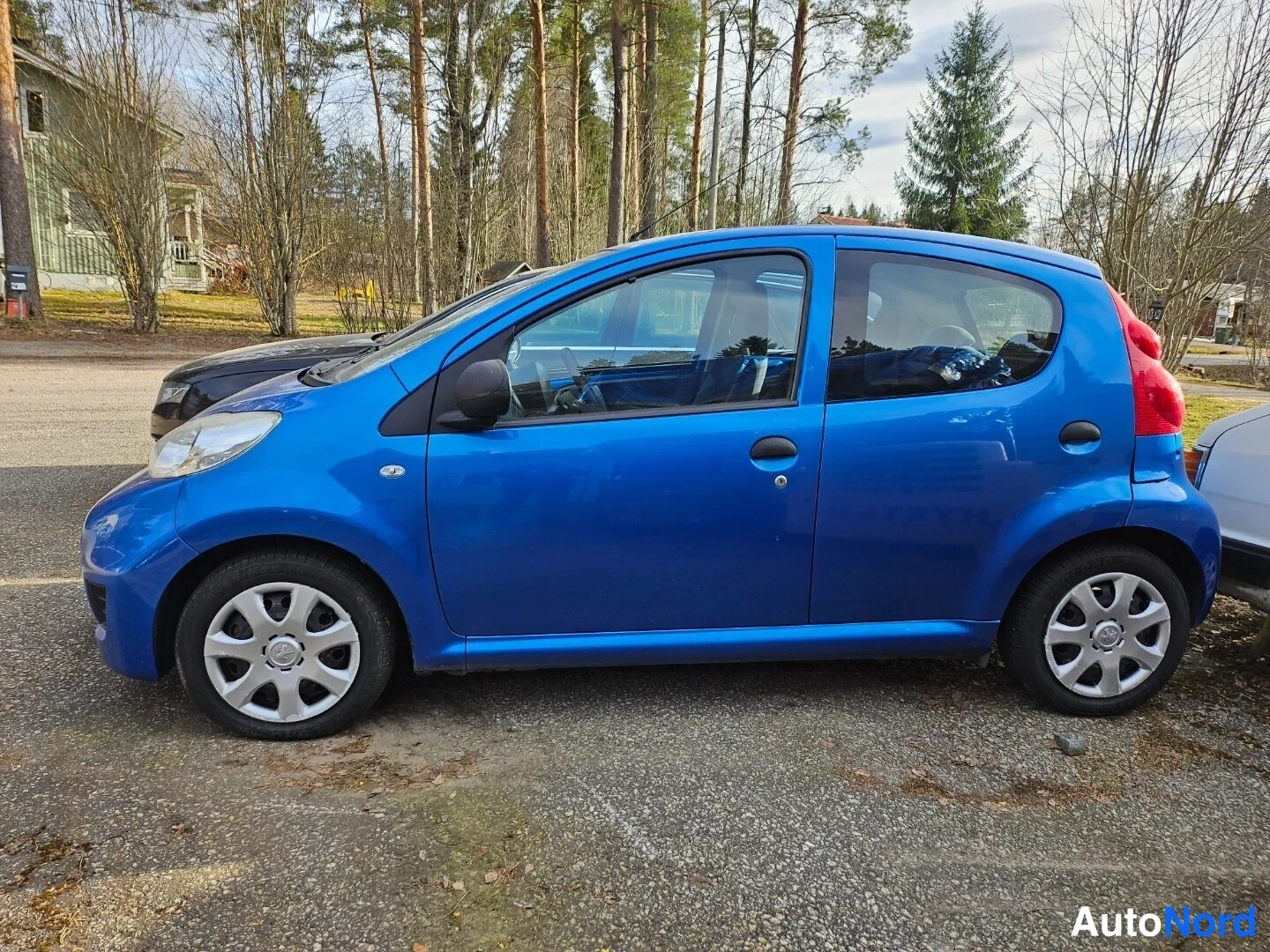 Sivulta: Sininen Peugeot 107 -henkilöauto sivulta kuvattuna, vasen kylki näkyvissä parkkipaikalla.