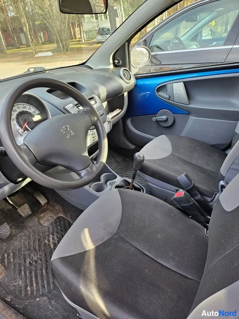 Edestä: Auton ohjaamo vasemmalta: Peugeot-ohjauspyörä, vaihtovipu, käsijarru ja tummat istuimet, sininen ovipaneeli oikealla.