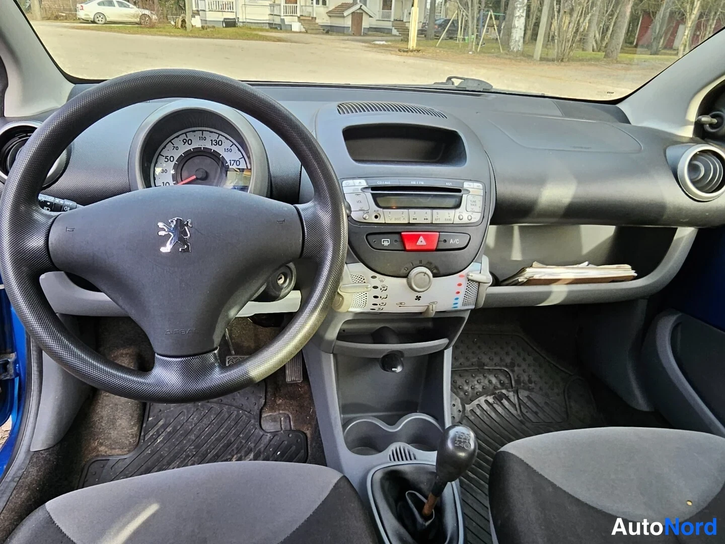 Edestä: Näkymä ohjaamoon: Peugeot-ohjauspyörä, nopeusmittari, keskikonsoli radio- ja A/C-painikkeilla, manuaalivaihteen vaihtovipu ja kumimatot.