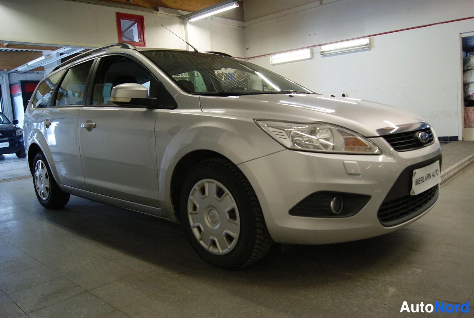 Etuviisto, nokka oikealle: Ford Focus Farmari 2008, hopeanvärinen, kulmakuva edestä oikealta korjaamotiloissa; etusäleikkö, ajovalot ja kylki näkyvissä.