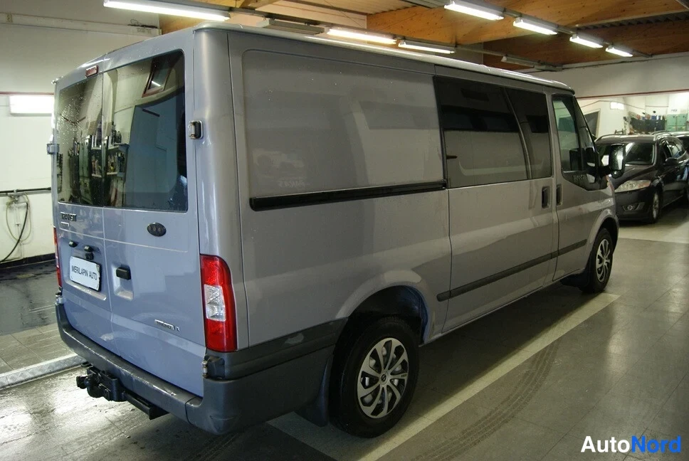 Takaviisto: Hopeanharmaa Ford Transit -pakettiauto takaa vasemmalta kuvattuna, perä- ja kylkiluukut auki, kori vaaleanharmaa hallissa.