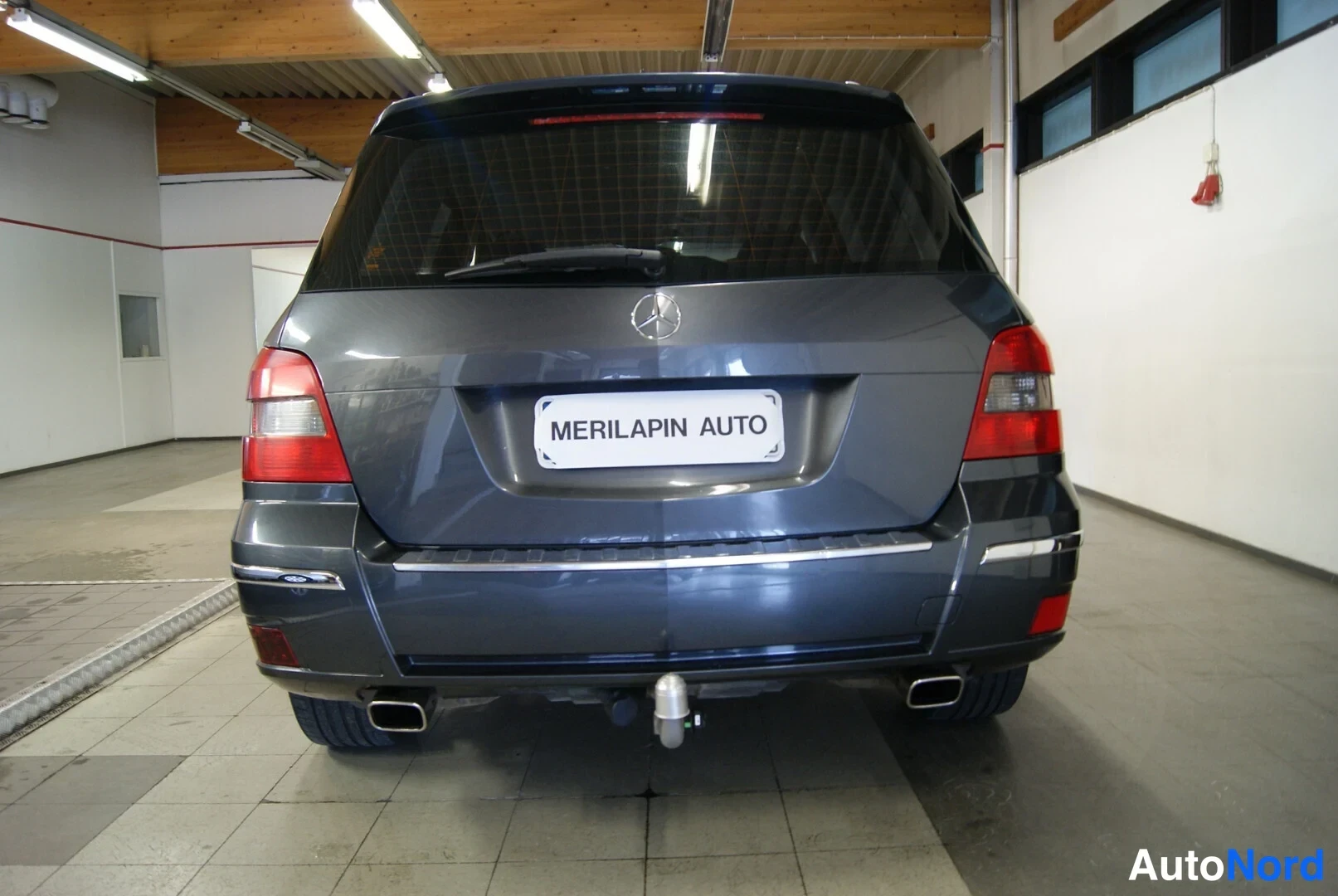 Takaa: Auton takanäkymä sisätilassa: tummanharmaa Mercedes-Benz, punaiset takavalot, vetokoukku ja kaksi pakoputkea.