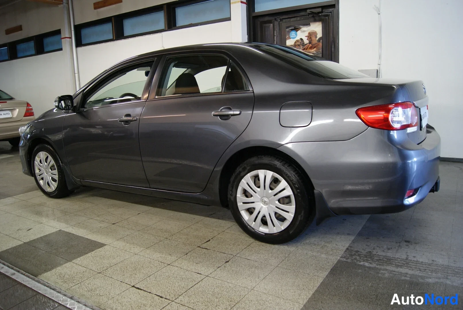 Takaviisto: Tummanharmaa Toyota Corolla -sedan takaa kolmesta neljänneksestä, takaosan ja kyljen näkyessä, autotallin sisätiloissa.