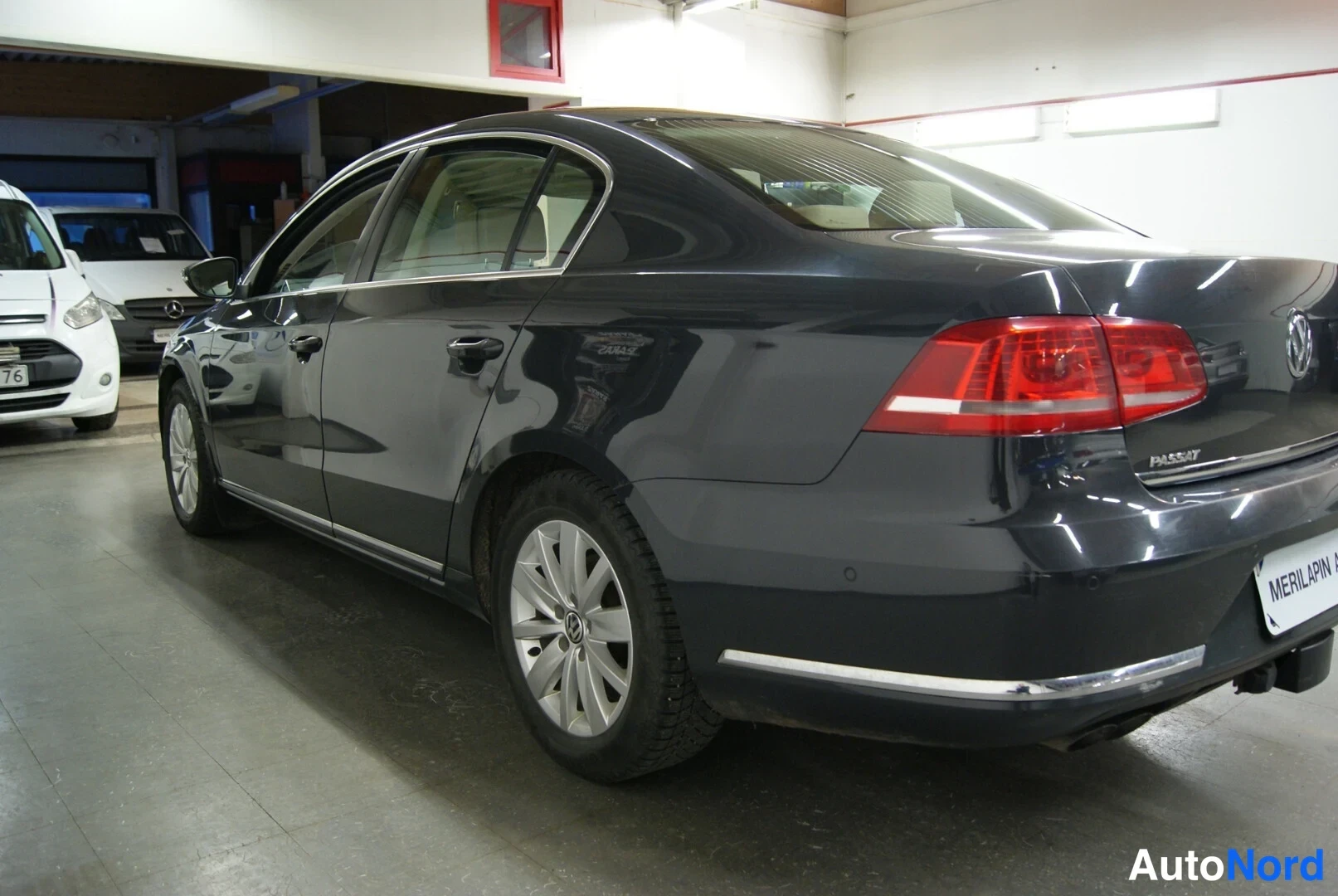 Takaviisto: Tummanharmaan Volkswagen Passat -sedanin takaosa ja vasen kylki myyntitilassa, takaluukussa PASSAT-teksti.