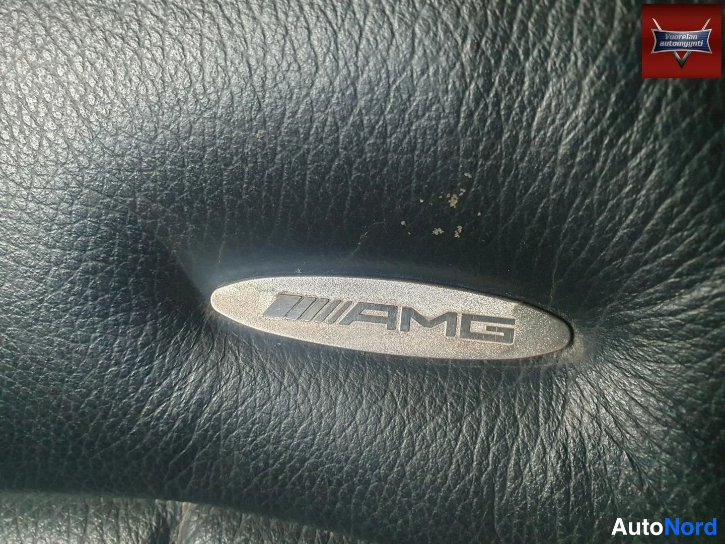 Yksityiskohta: Lähikuva mustan nahkapinnan keskellä olevasta metallisesta AMG-logokyltistä.