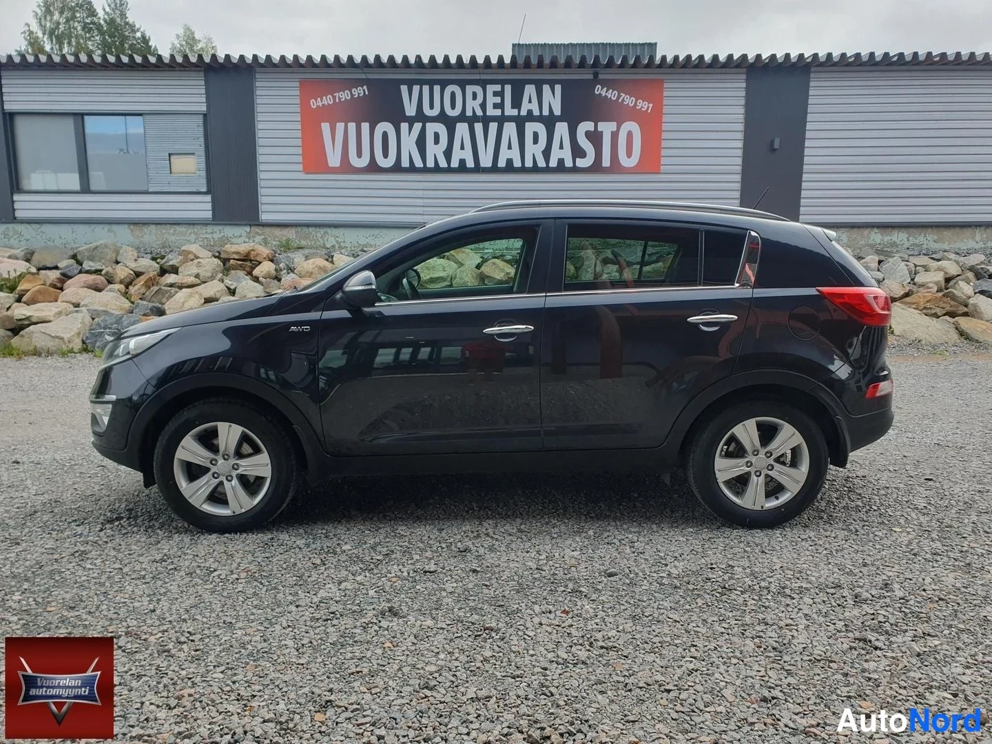 Sivulta: Musta Kia Sportage -mallinen maastokuja sivulta kuvattuna sorapitoisella pihalla, taustalla Vuorelan Vuokravarasto-talo.