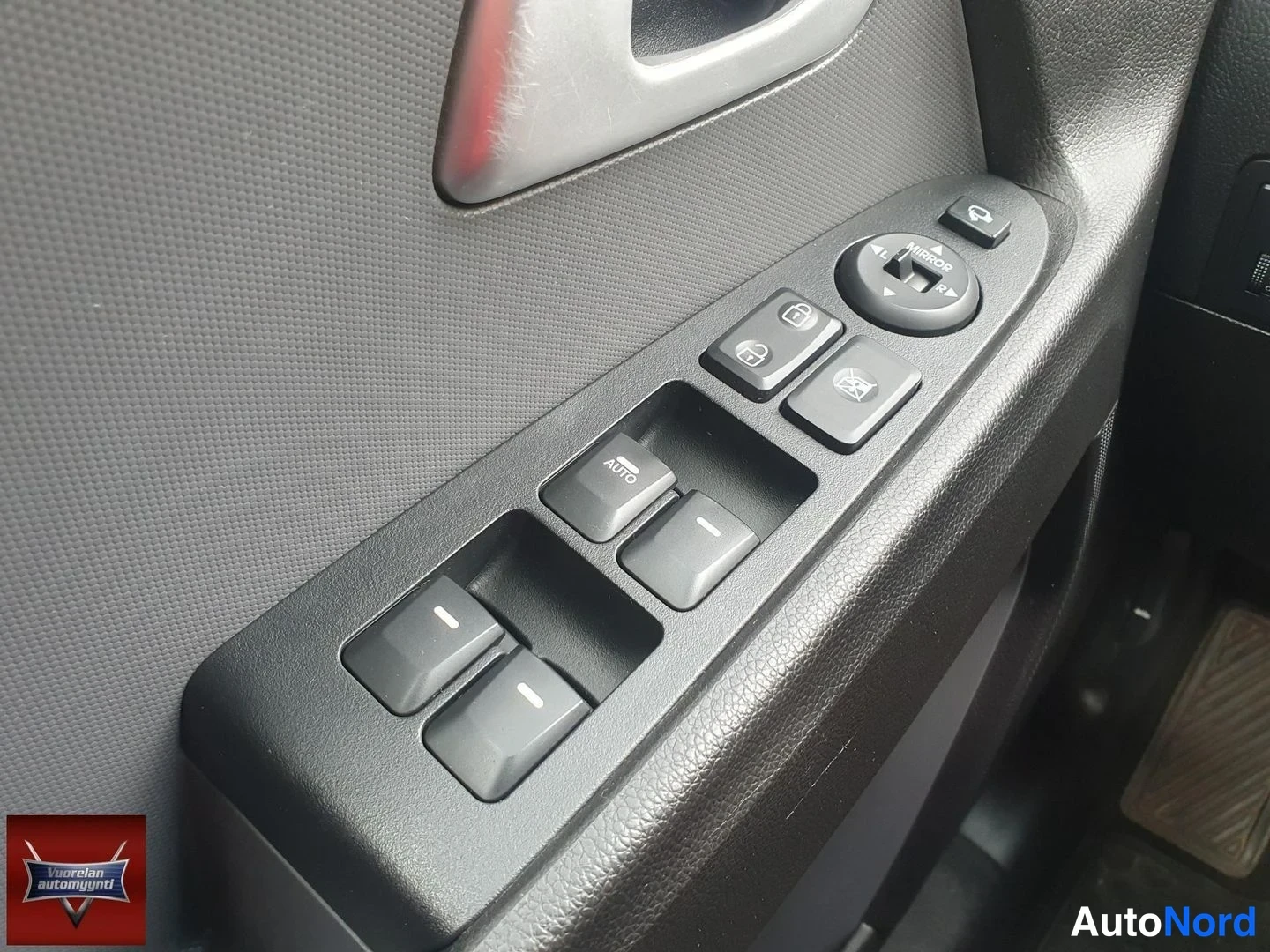 Yksityiskohta: Auton oven sisäpaneelin kytkinryhmä lähikuvassa: sähköikkunoiden painikkeet (toisessa teksti AUTO), peilien säätökiekko MIRROR-merkinnällä sekä lukitus- ja ikku