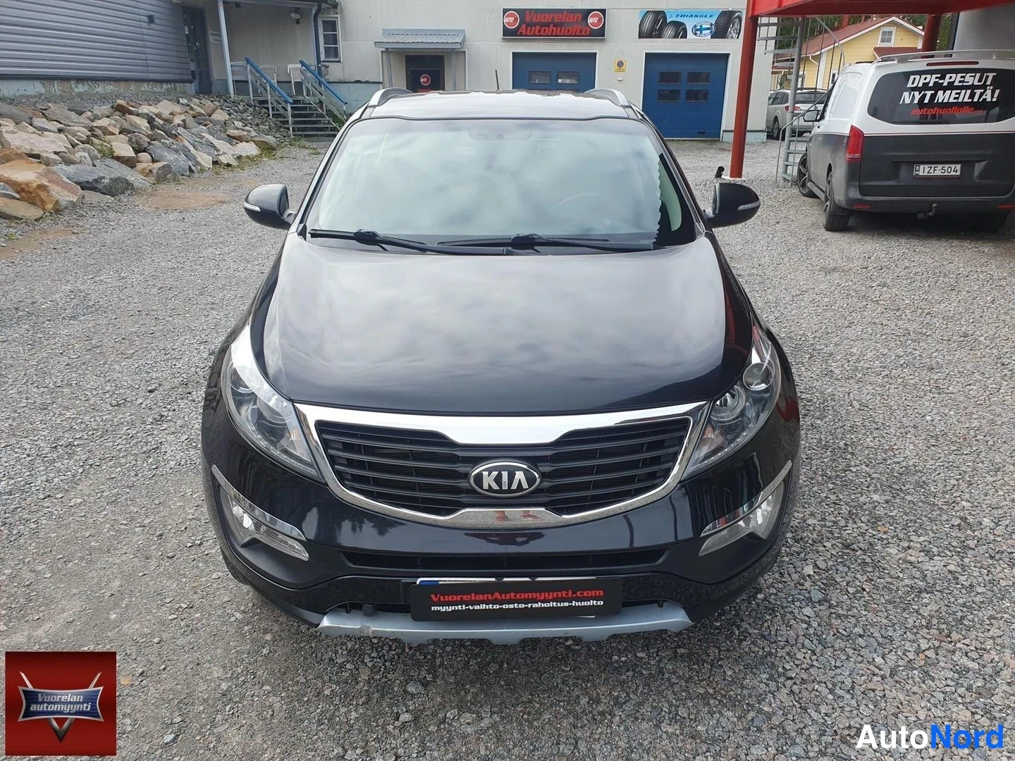 Edestä: Suoraan edestä kuvattu musta Kia Sportage, KIA-merkki ja etusumuvalot näkyvät, taustalla autokorjaamon siniset bayt.