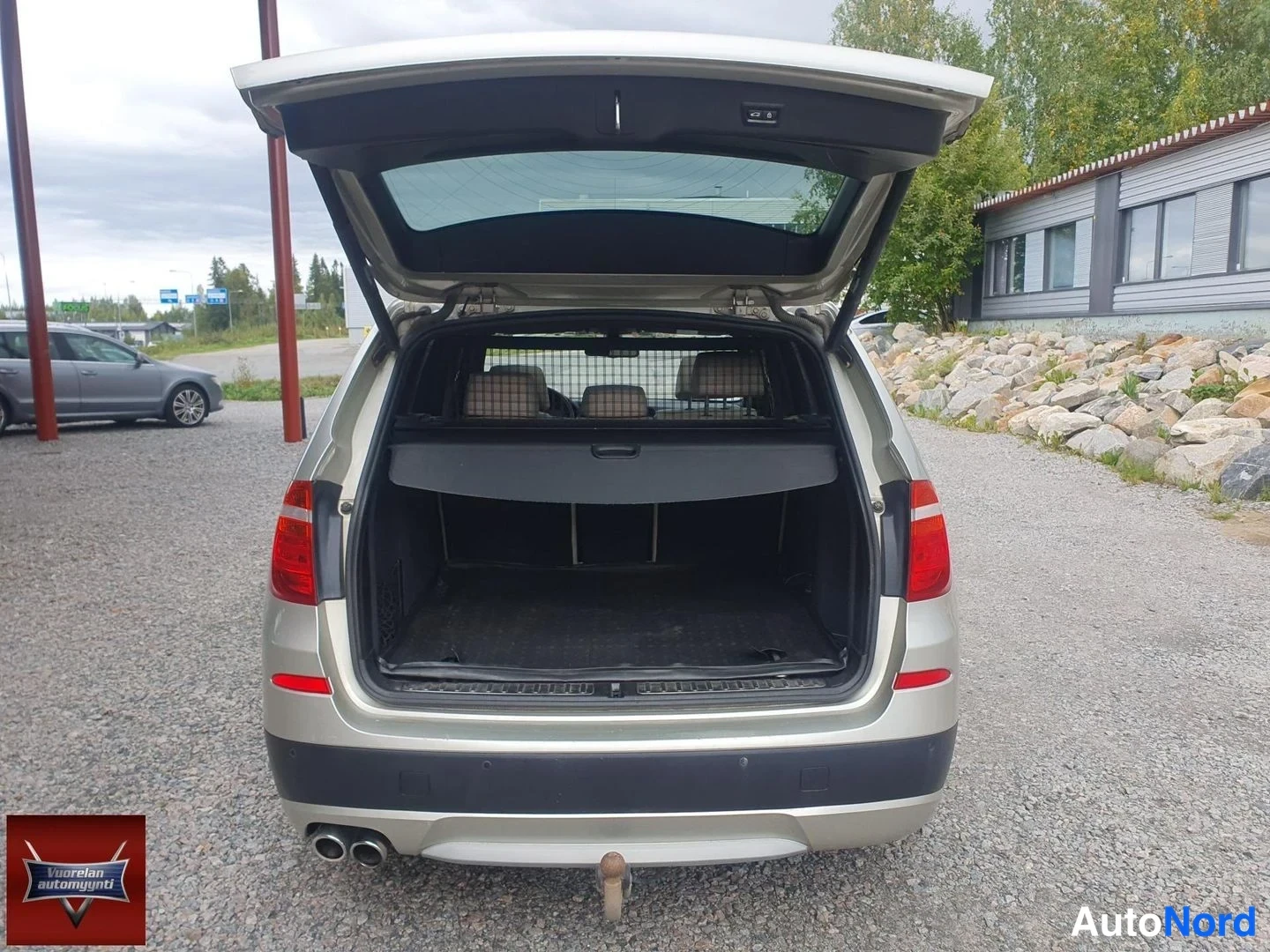 Takaa: Takaa suoraan kuvattu auto, takaluukku auki: tavaratilassa näkyy tumma matto ja tavaratilan suojaverkko, takapuskurissa kaksi pakoputkea ja vetokoukun pallo.