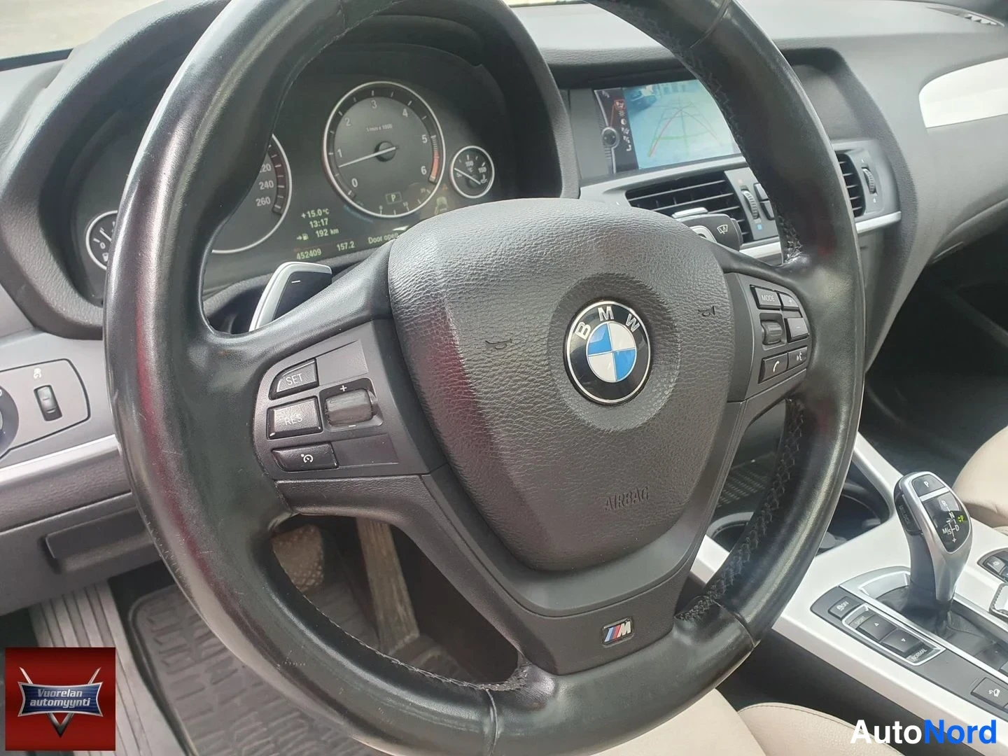 Muu: Ohjauspyörä BMW-embleemillä ja M-merkinnällä, mittaristo taustalla, navigointinäyttö ja vaihteenvalitsin osin näkyvissä.
