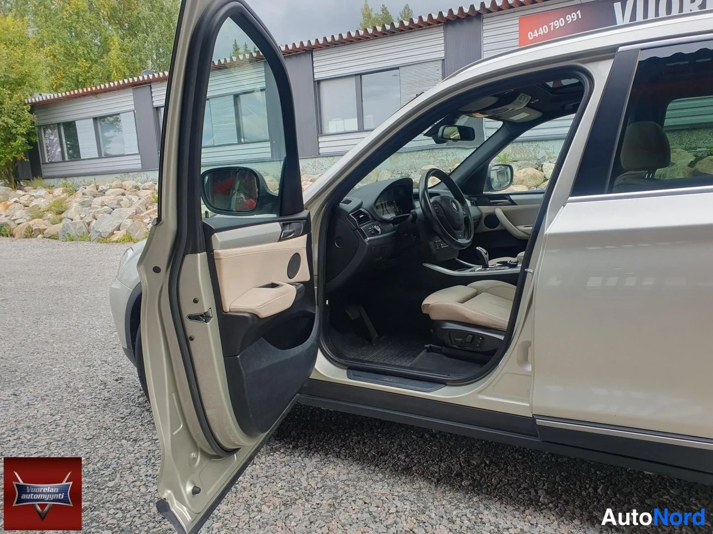 Sivulta: Sivukuva BMW X3:sta, kuljettajan puolen etuovi auki. Näkyvissä vaaleanbeige nahkaverhoiltu sisusta: ohjauspyörä, kuljettajan istuin ja vaihteenvalitsin.