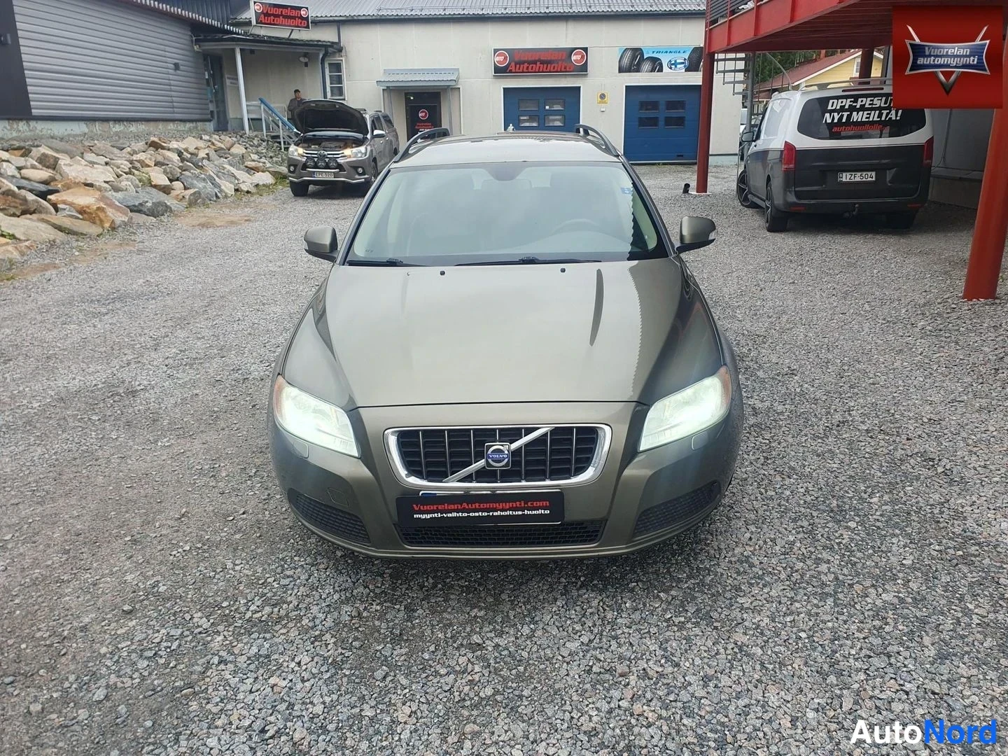 Edestä: Oliivinruskeanharmaa Volvo V70 -farmari kuvattuna suoraan edestä sorapihalla. Etusäleikössä Volvon merkki, ja etupuskurin alapuolella liikkeen mainoskyltti. Tau