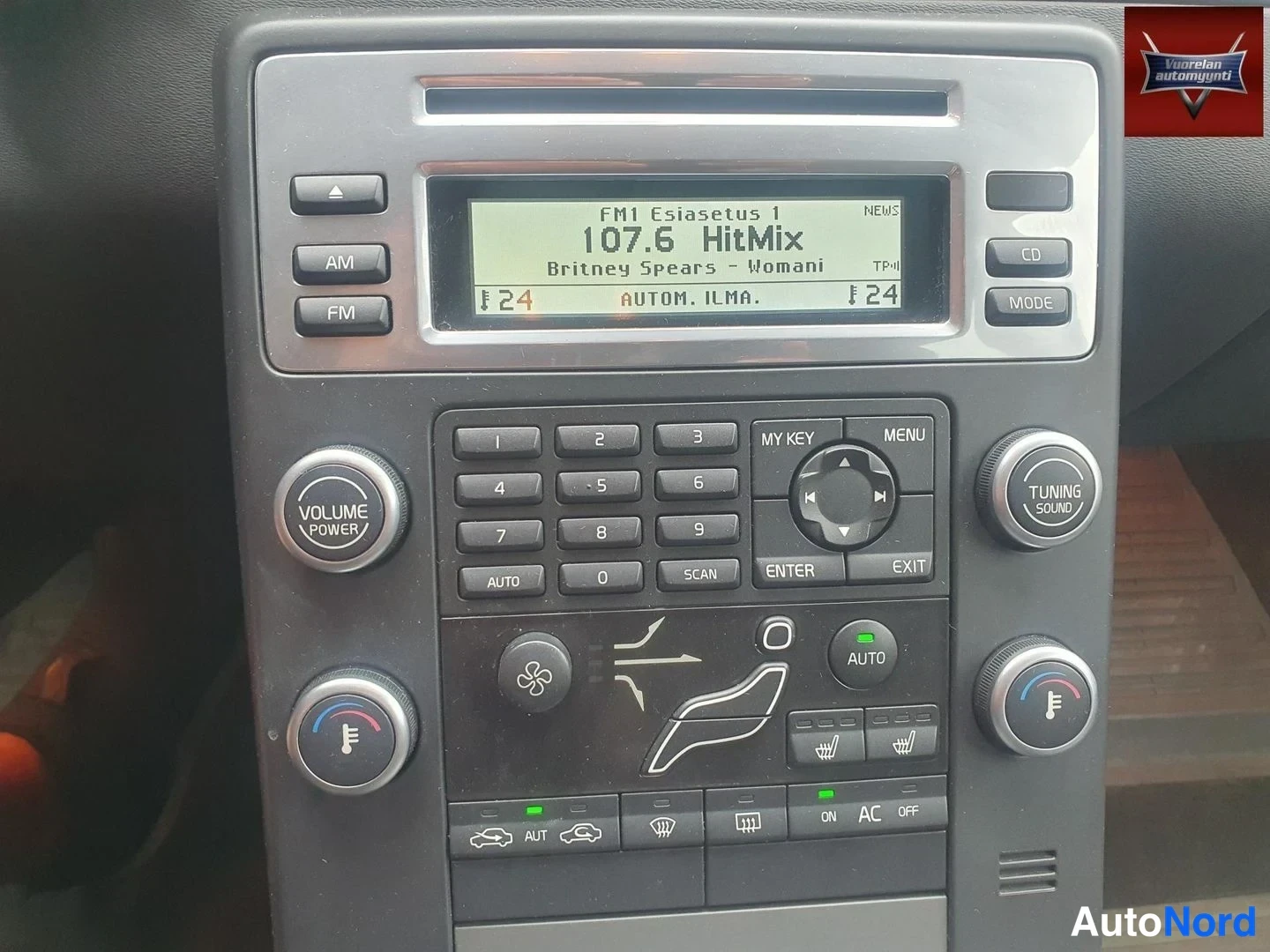 Yksityiskohta: Autojen keskikonsolin lähikuva: radio-CD-paneeli ja automaattinen ilmastointi, näytöllä lukee 'FM1 107.6 HitMix' ja 'AUTOM. ILMA.'.