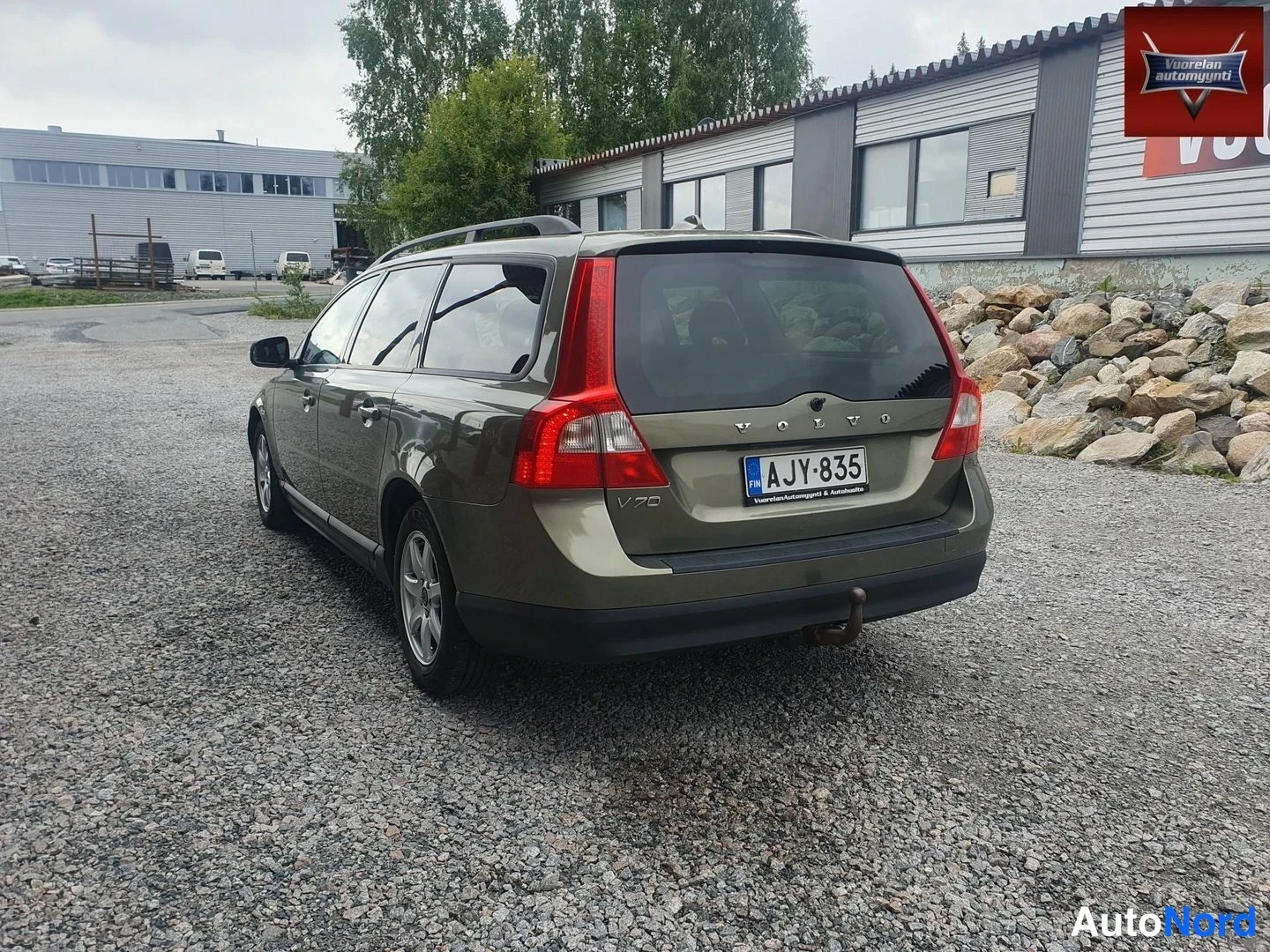 Takaviisto: Oliivinvihreä Volvo V70 -farmuri takaa viistosti, rekisterikilpi, vetokoukku ja kattokaiteet näkyvissä, sorapiha ja rakennuksia taustalla.
