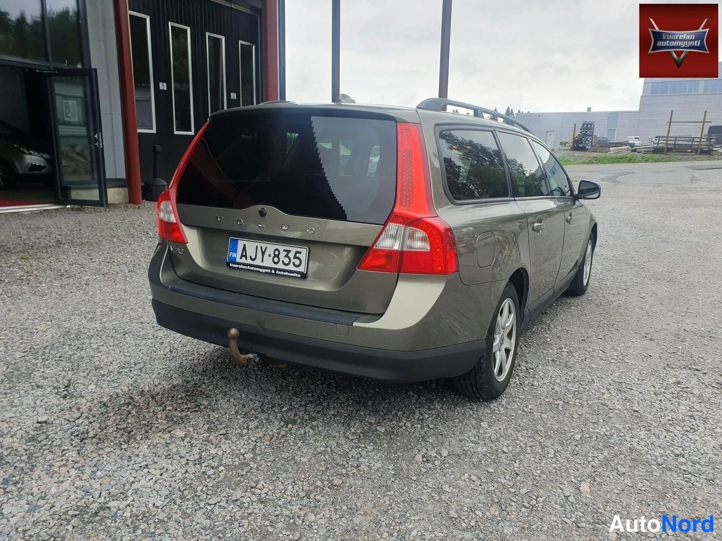 Takaviisto: Oliivinvihreä Volvo V70 -farmuri takaa viistosti, rekisterikilpi, vetokoukku ja kattokaiteet näkyvissä, sorapiha ja rakennuksia taustalla.