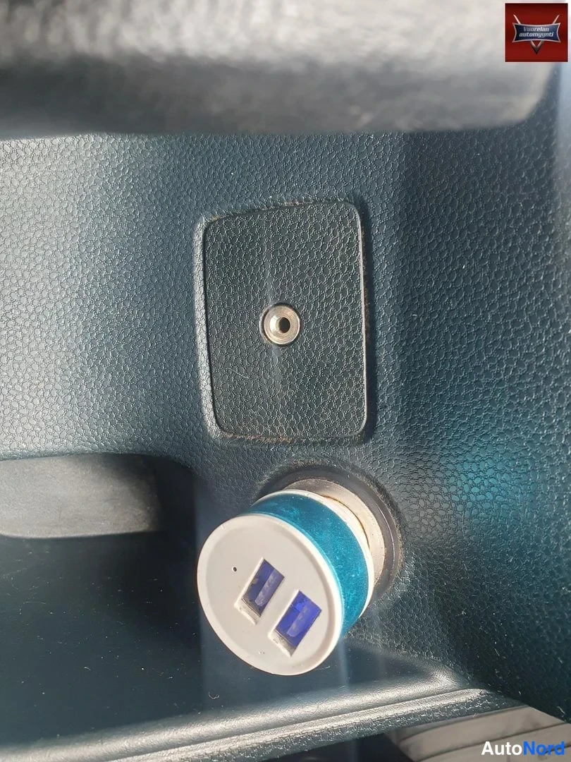 Yksityiskohta: Sisätilan lähikuva: tummassa teksturoidussa kojelautamuovissa olevaan 12 V -pistorasiaan on kytketty kaksois-USB-adapteri, jonka kaksi sinistä USB-porttia näkyv