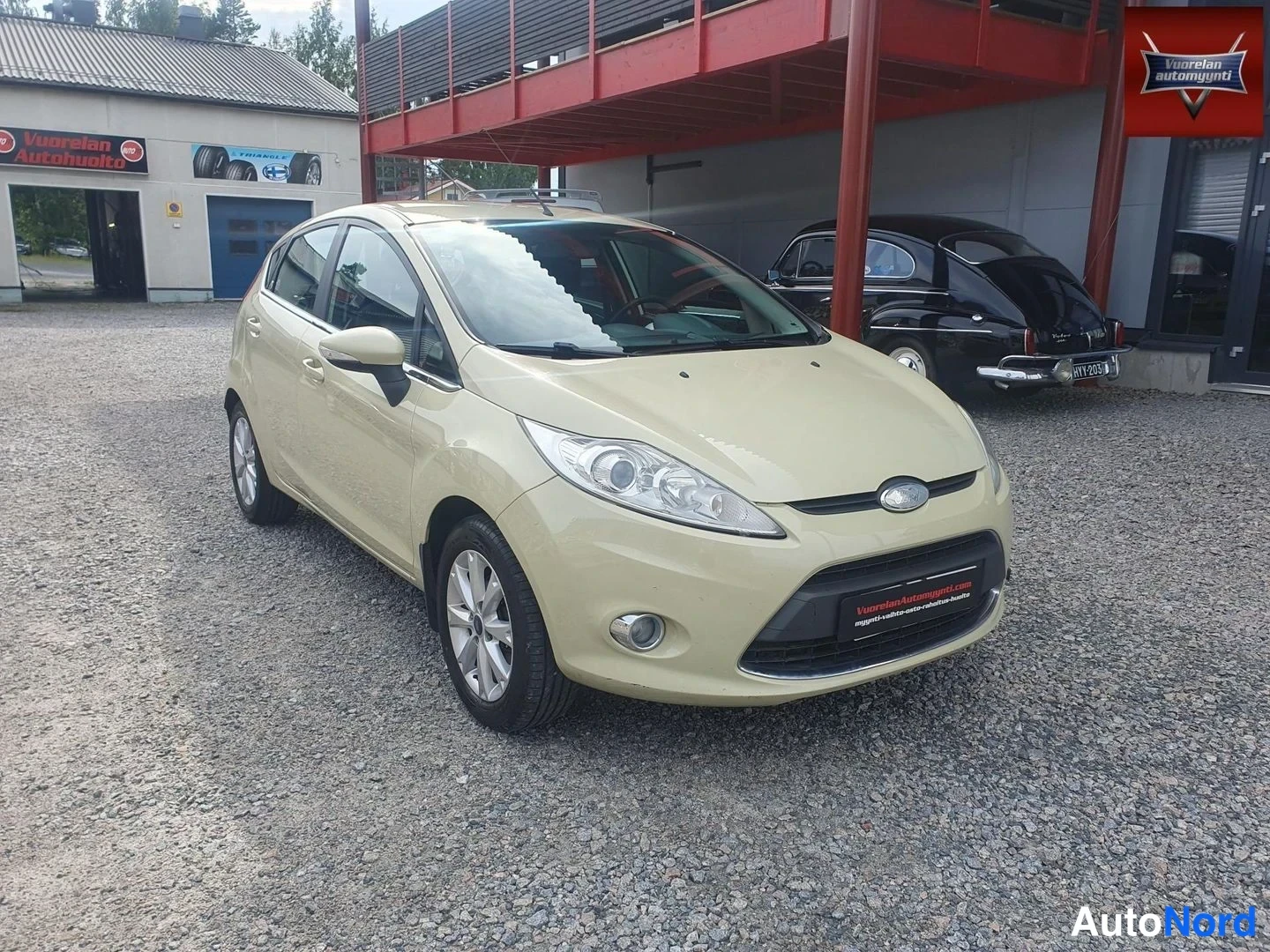 Etuviisto, nokka oikealle: Beige Ford Fiesta -henkilöauto kuvattuna etuviistosta: etusäleikkö, ajovalot ja etupuskuri kuvan oikeassa laidassa, auton vasen kylki ja takapyhä näkyvis