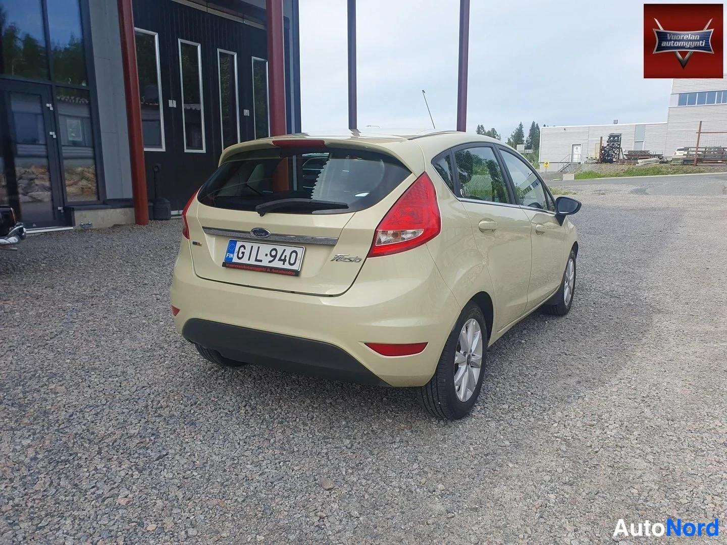 Takaviisto: Beige Ford Fiesta takaviistosta: takavalot, takaluukku ja rekisterikilpi näkyvissä, auton oikea kylki kuvan oikealla puolella.