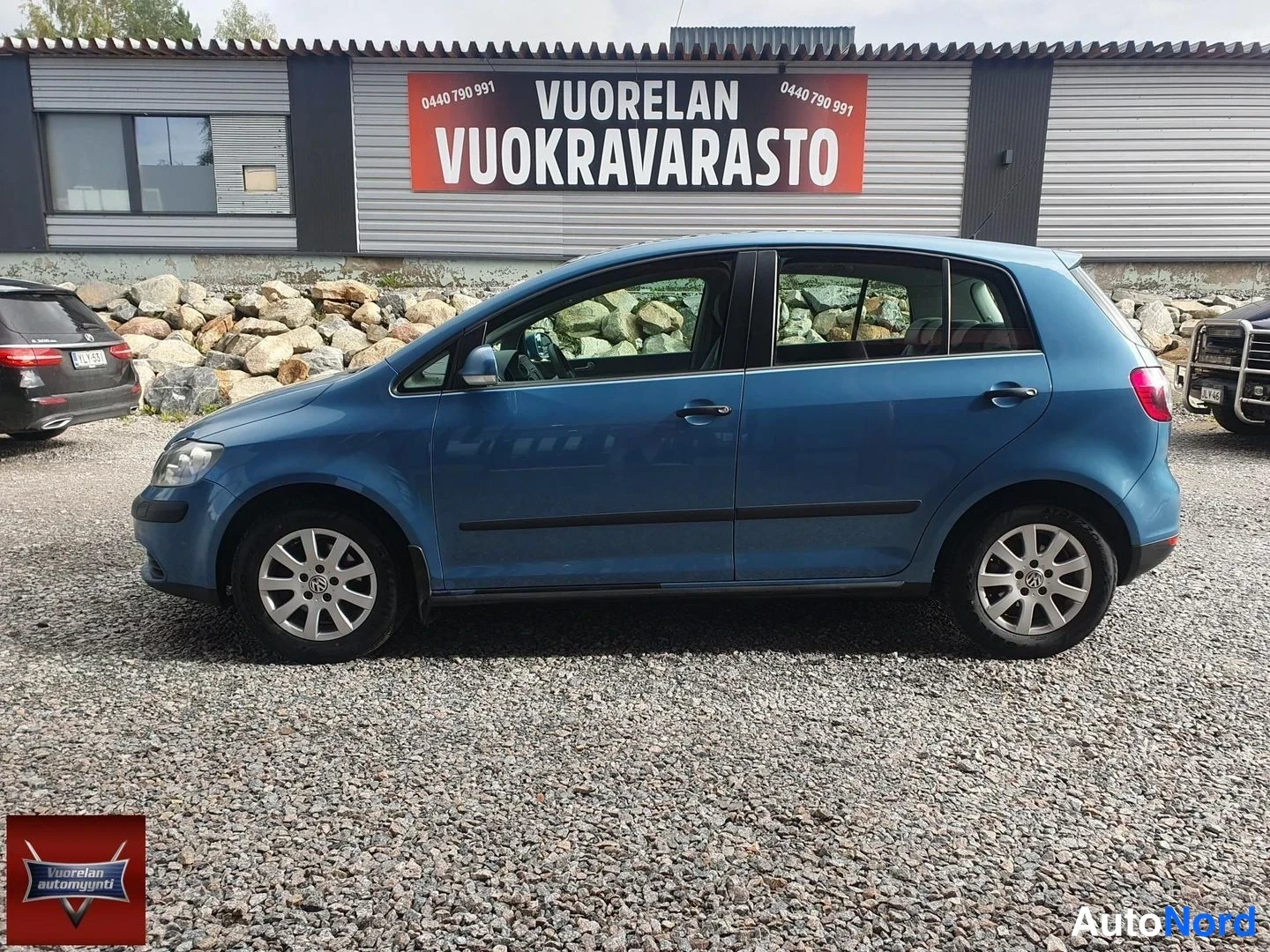 Sivulta: Sininen henkilöauto kuvattuna sivulta hieman edestä, keula vasemmalla, sorapihalla Vuorelan Vuokravarasto-rakennuksen edessä.