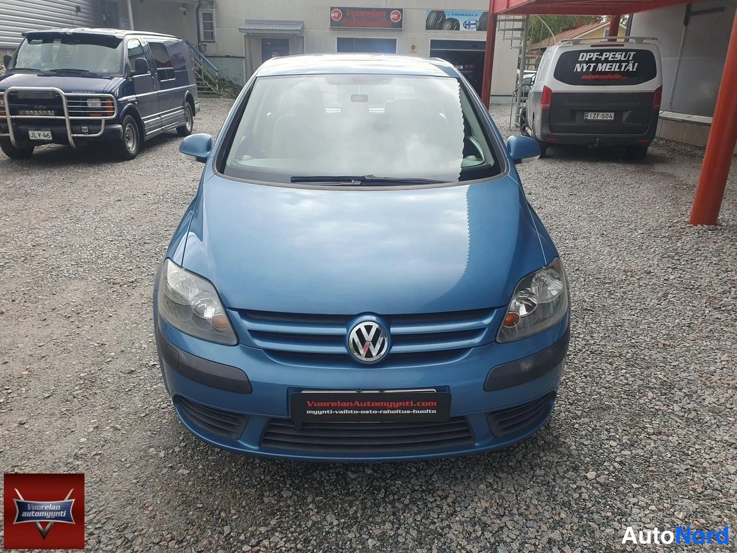 Edestä: Sininen Volkswagen Golf Plus kuvattuna suoraan edestä: etusäleikkö VW-merkillä, ajovalot ja etupuskuri näkyvät, taustalla sorapiha ja muita ajoneuvoja.